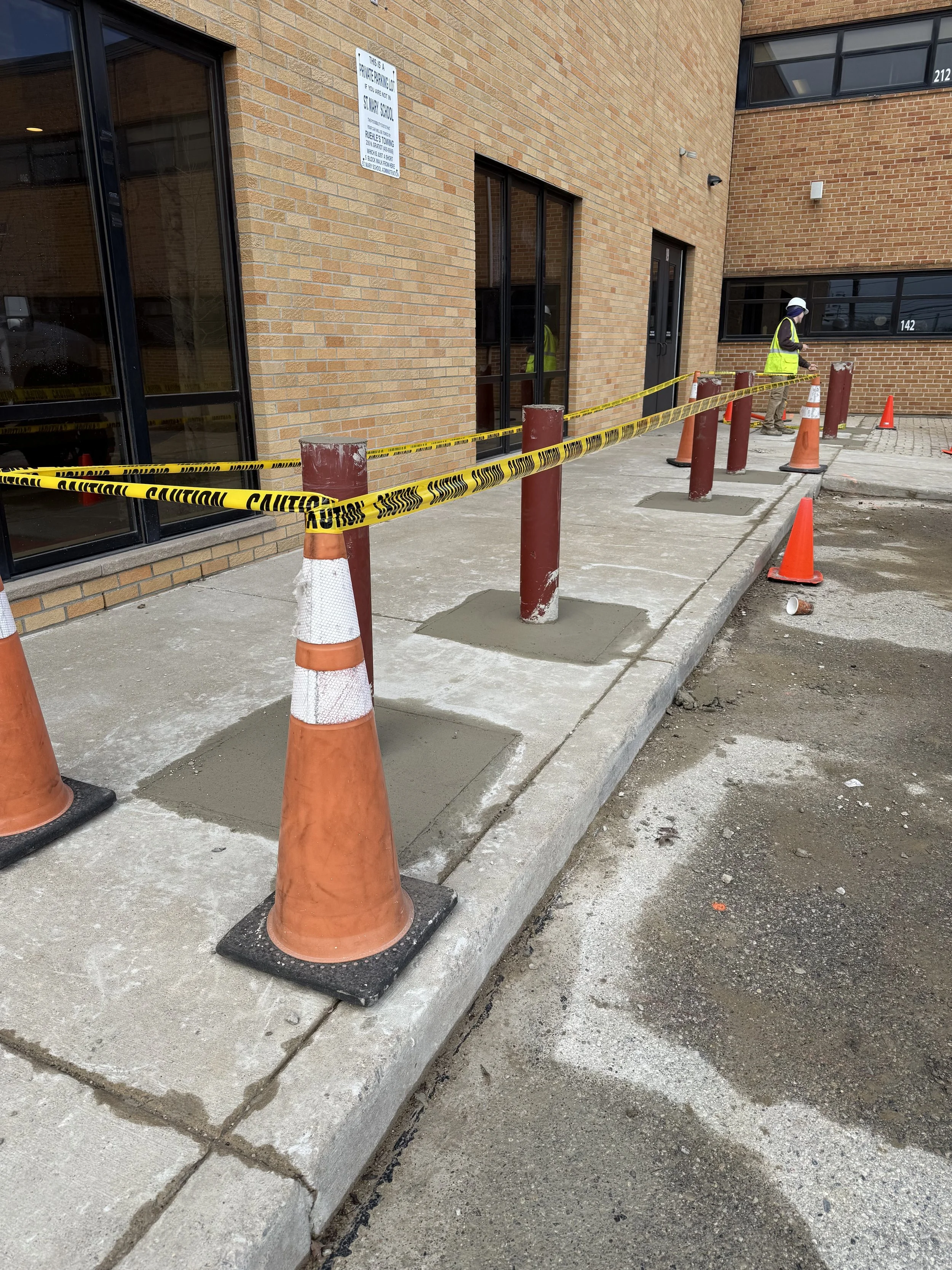 ASTM F2656 crash-rated bollards in primer coat set in fresh concrete along main entrance walkway at St. Mary Catholic School, Mount Clemens Michigan, during NSGP-funded installation by Trident Industrial Systems