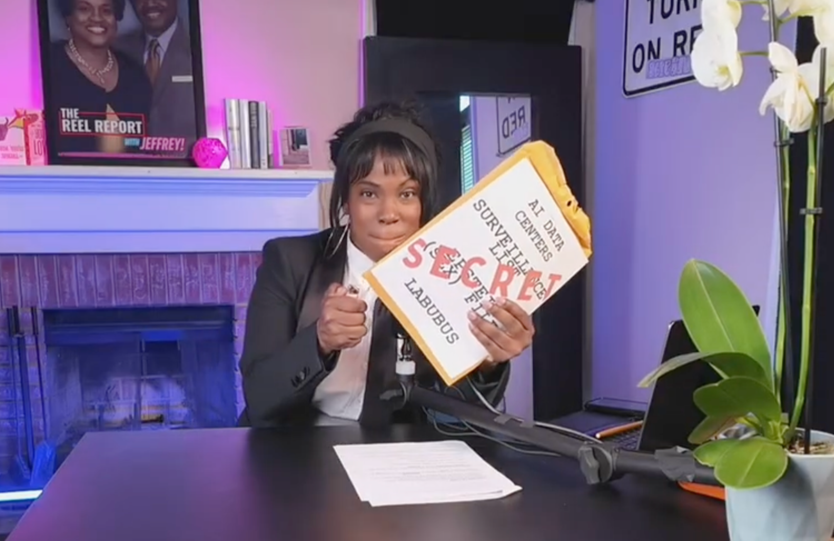 A woman in a black blazer and white shirt holding a sign that says 'AT DARY CENTER SURVIVORS LIKE THE SEX RESEARCH LABORATORY' while sitting at a desk with a microphone, a sheet of paper, and a green plant in a white pot.