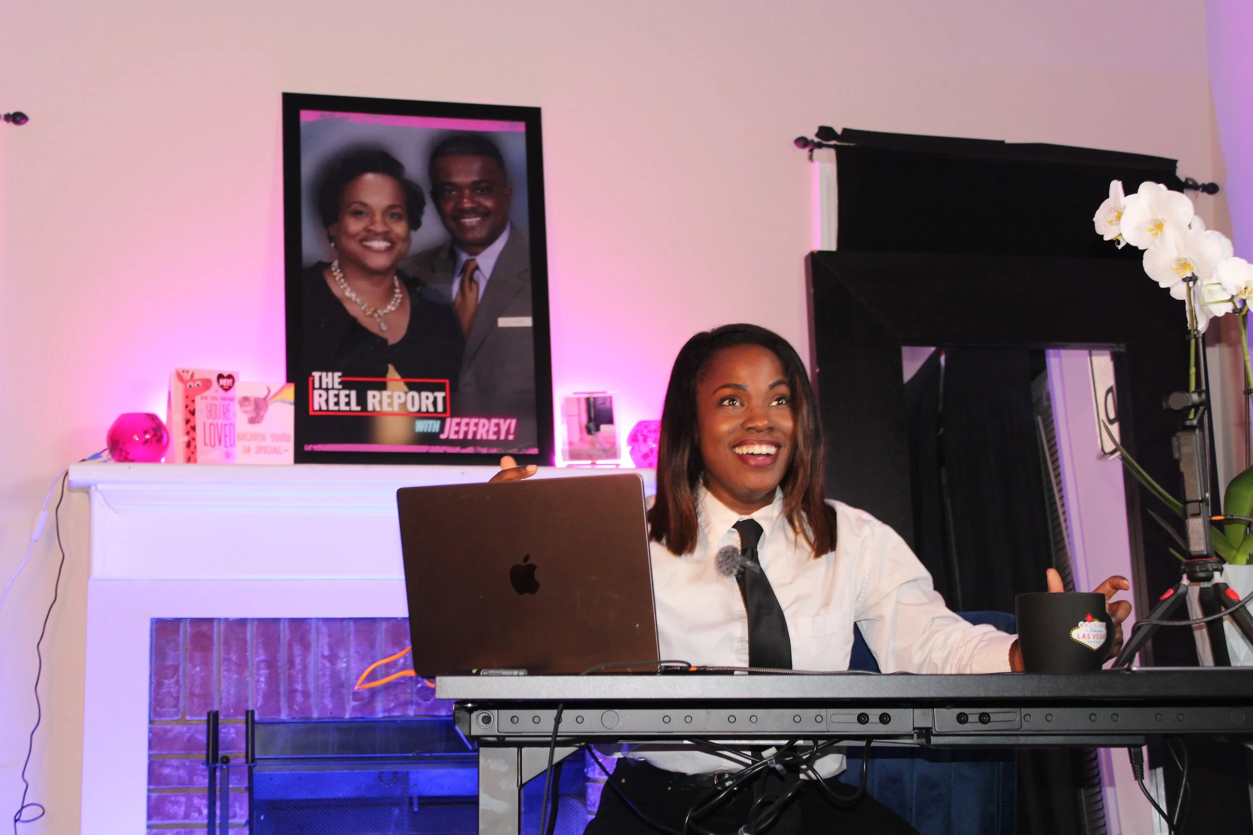 A woman sitting at a desk in front of a laptop, smiling, with a professional photo on the wall behind her showing a man and woman smiling, both dressed formally. The desk has a microphone, a black cup with a Las Vegas emblem, and other items. There are pink and purple lighting accents around the room.