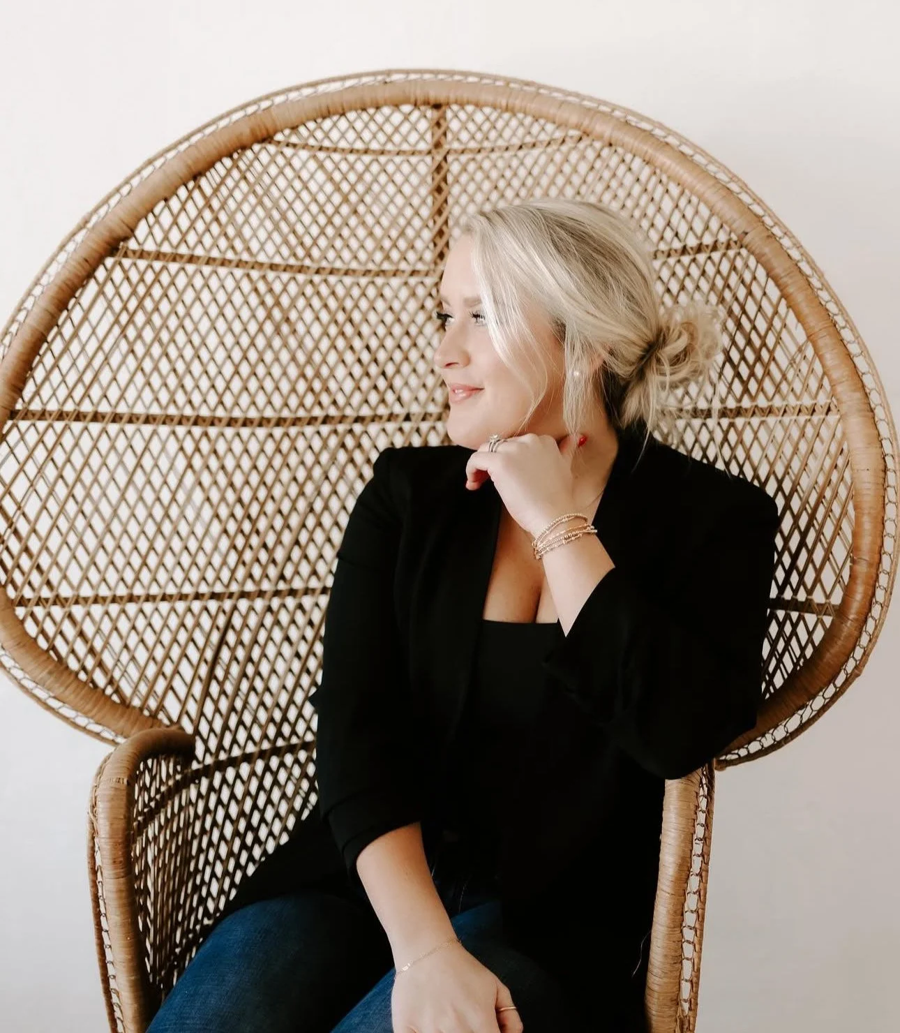 A woman with blonde hair tied back, wearing a black blazer, sitting in a large wicker peacock chair, smiling while looking to the side.