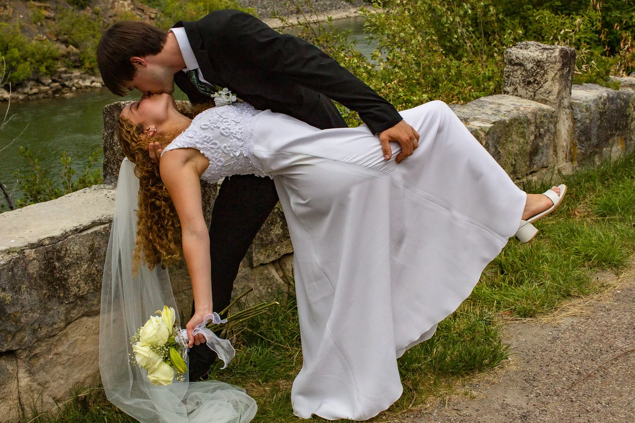 A bride and groom sharing a romantic kiss outdoors near a stone wall and water.
