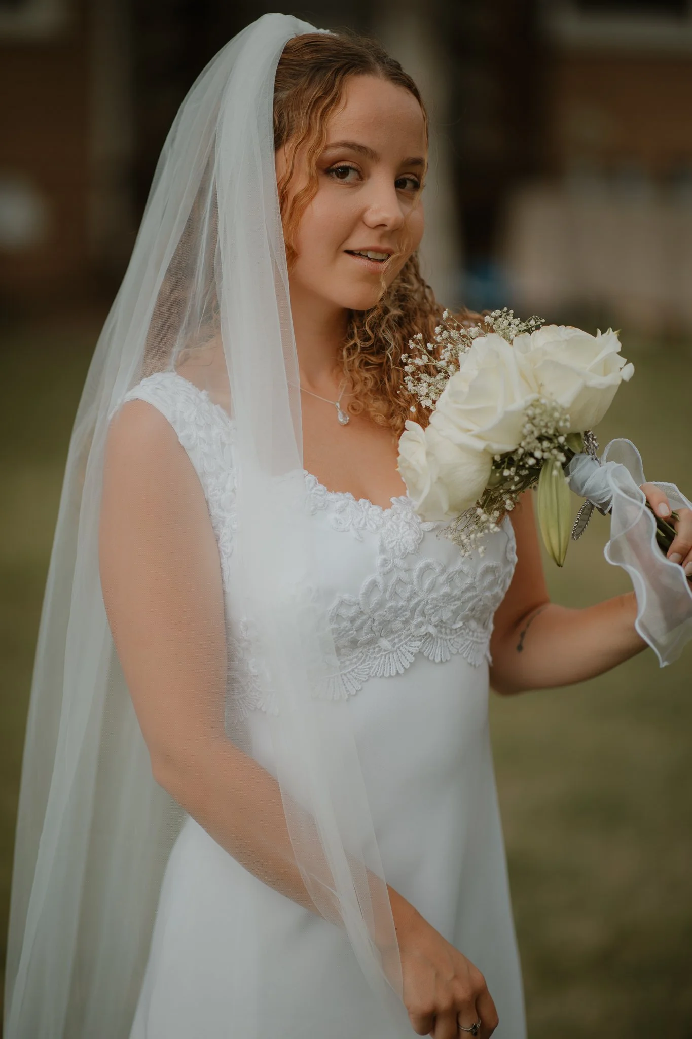 A woman in a white wedding dress and veil holding a bouquet of white roses, standing outdoors.