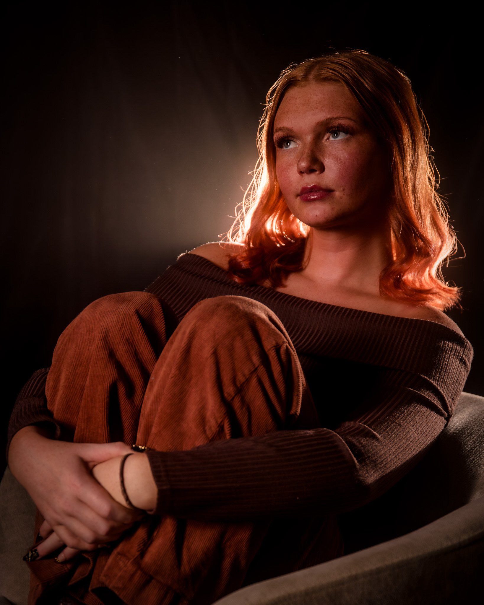 A young woman with red hair sitting on a sofa, looking thoughtful, with a dark background and backlighting highlighting her profile.