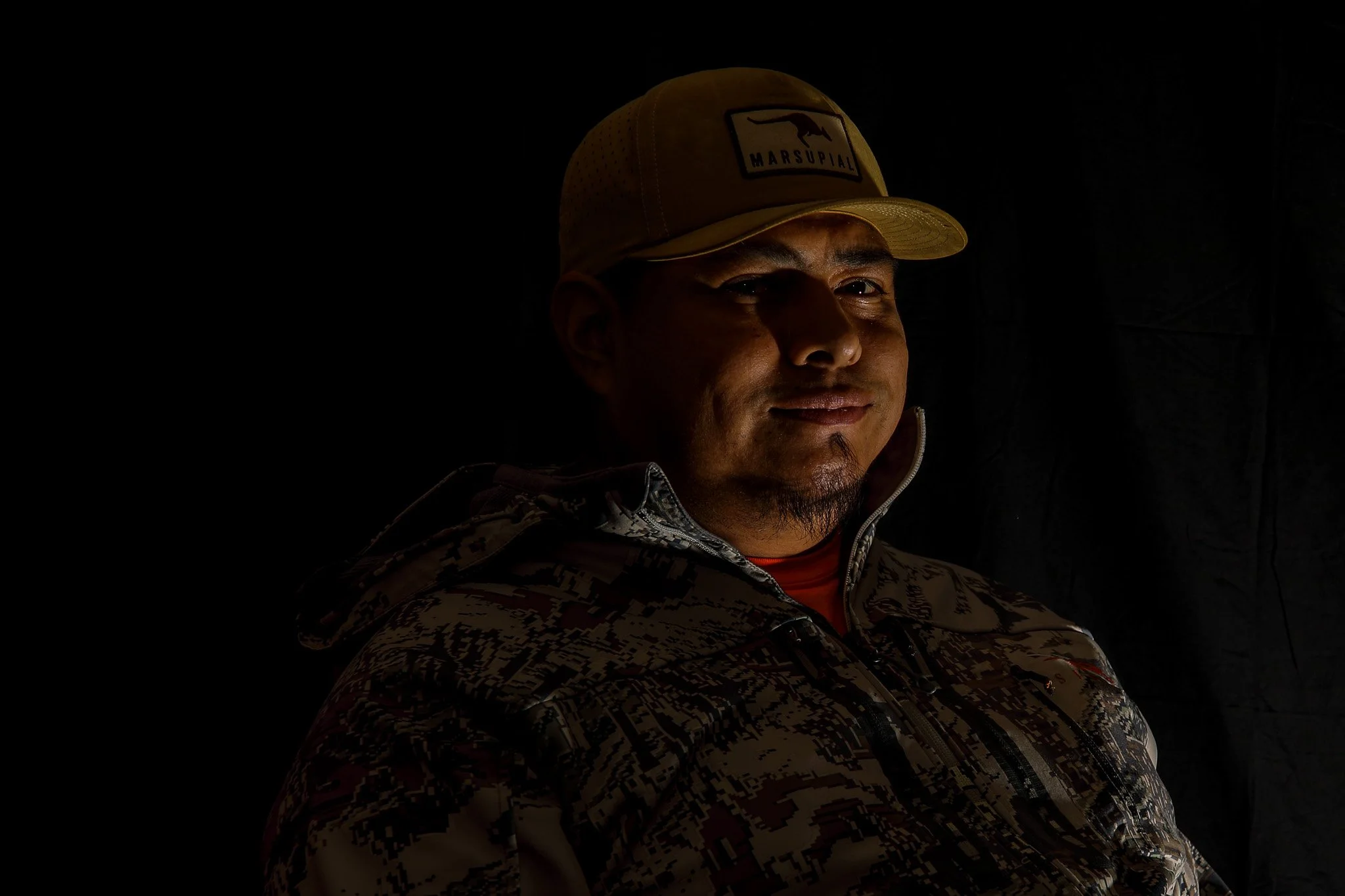 Portrait of a man wearing a beige cap with a dog and Marsupial logo and a camouflage jacket, illuminated from the side against a dark background.