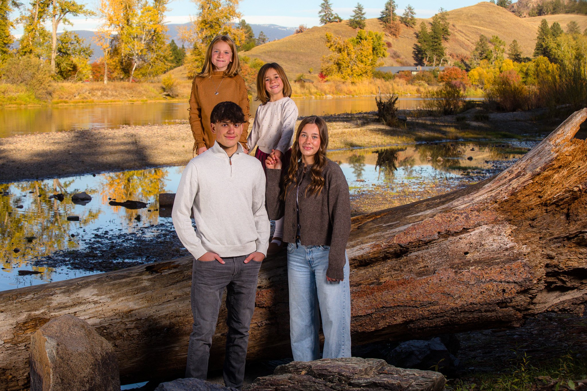 Four children, two boys and two girls, stand outdoors near a fallen tree by a river, with autumn-colored trees and hills in the background.