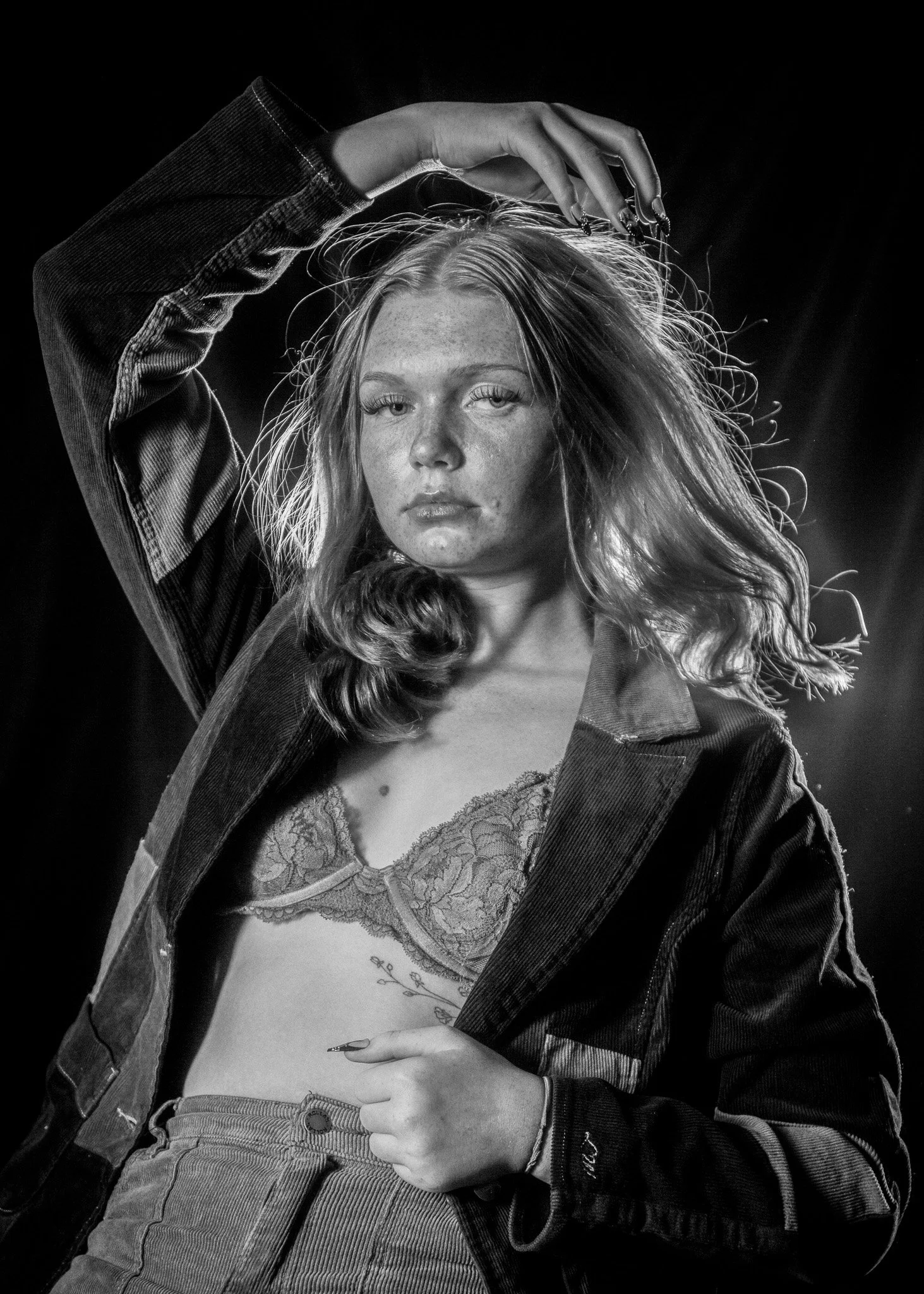 Black and white photograph of a young woman with wavy hair, wearing a lace top, denim jacket, and high-waisted pants, posing with her hand near her head against a dark background.