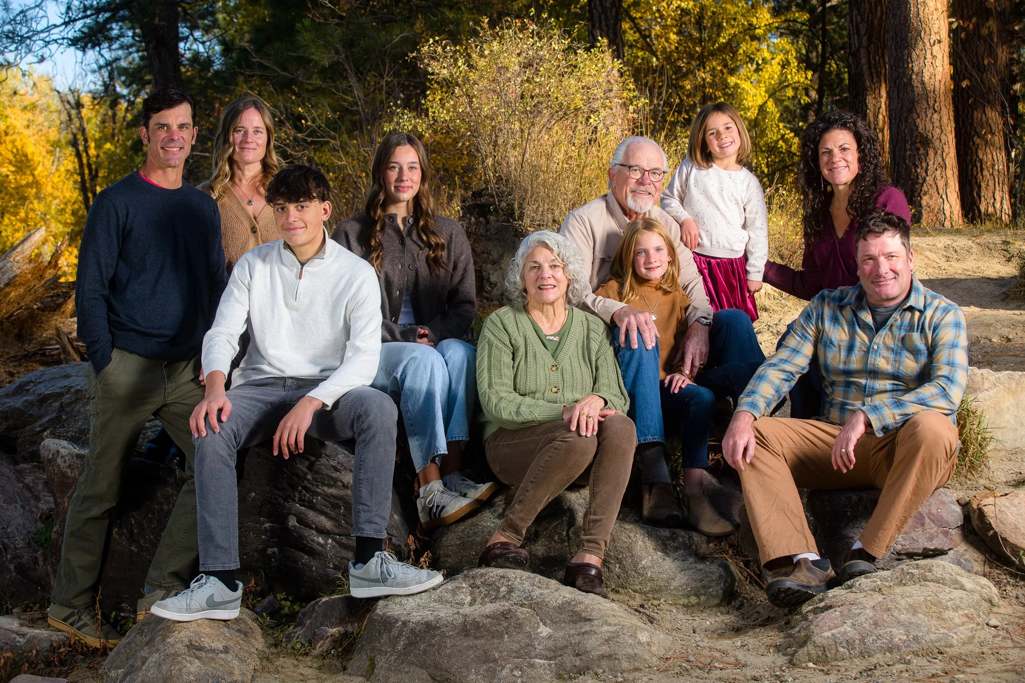 A large family outdoors in a wooded area with autumn foliage, posing on rocks for a group photo. They include two elderly individuals, adults, teenagers, and young children, all dressed casually.