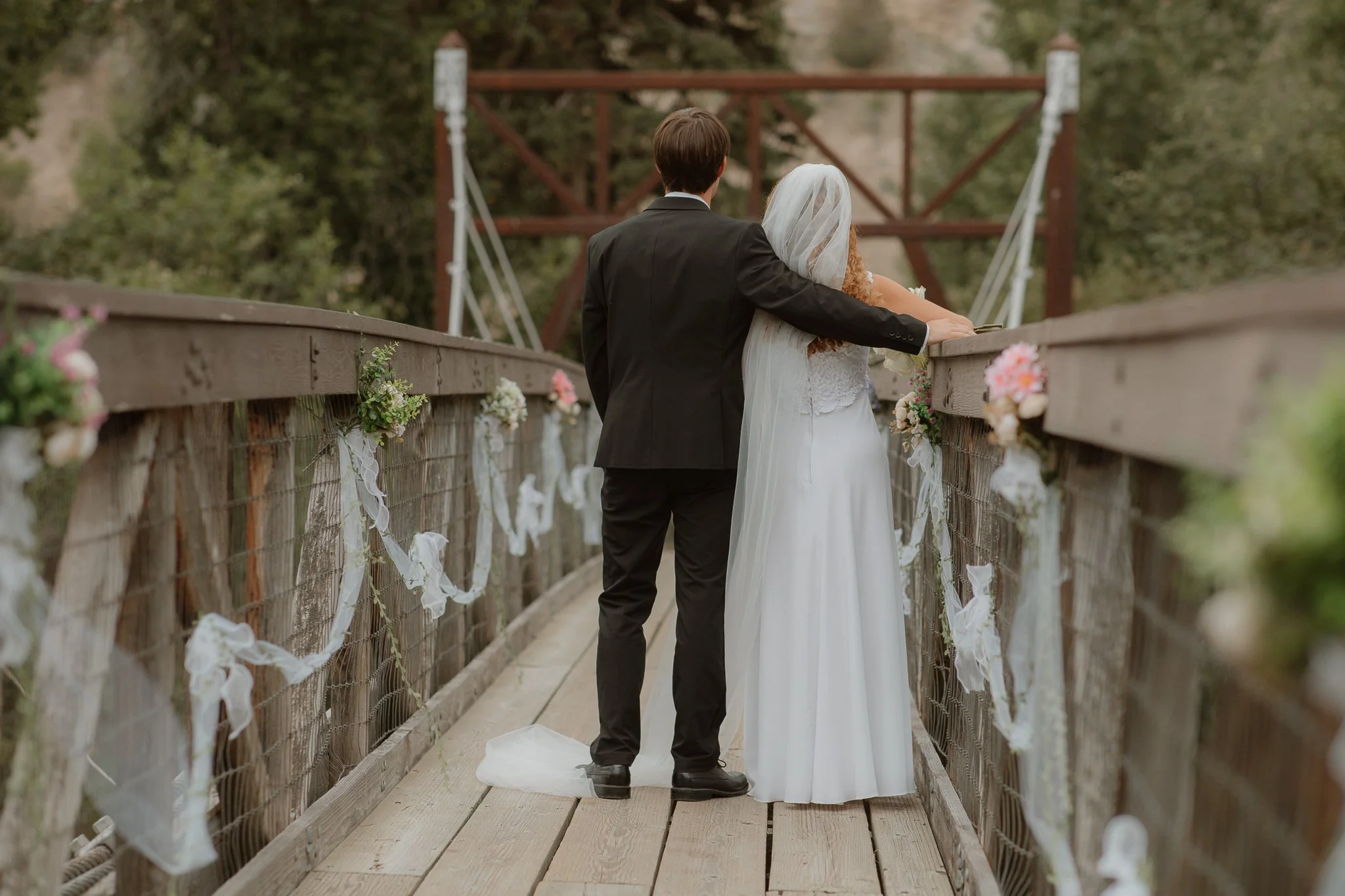 A bride and groom standing on a decorated wooden bridge, facing away, with the groom's arm around the bride, overlooking a natural landscape with trees and a bridge structure in the background.