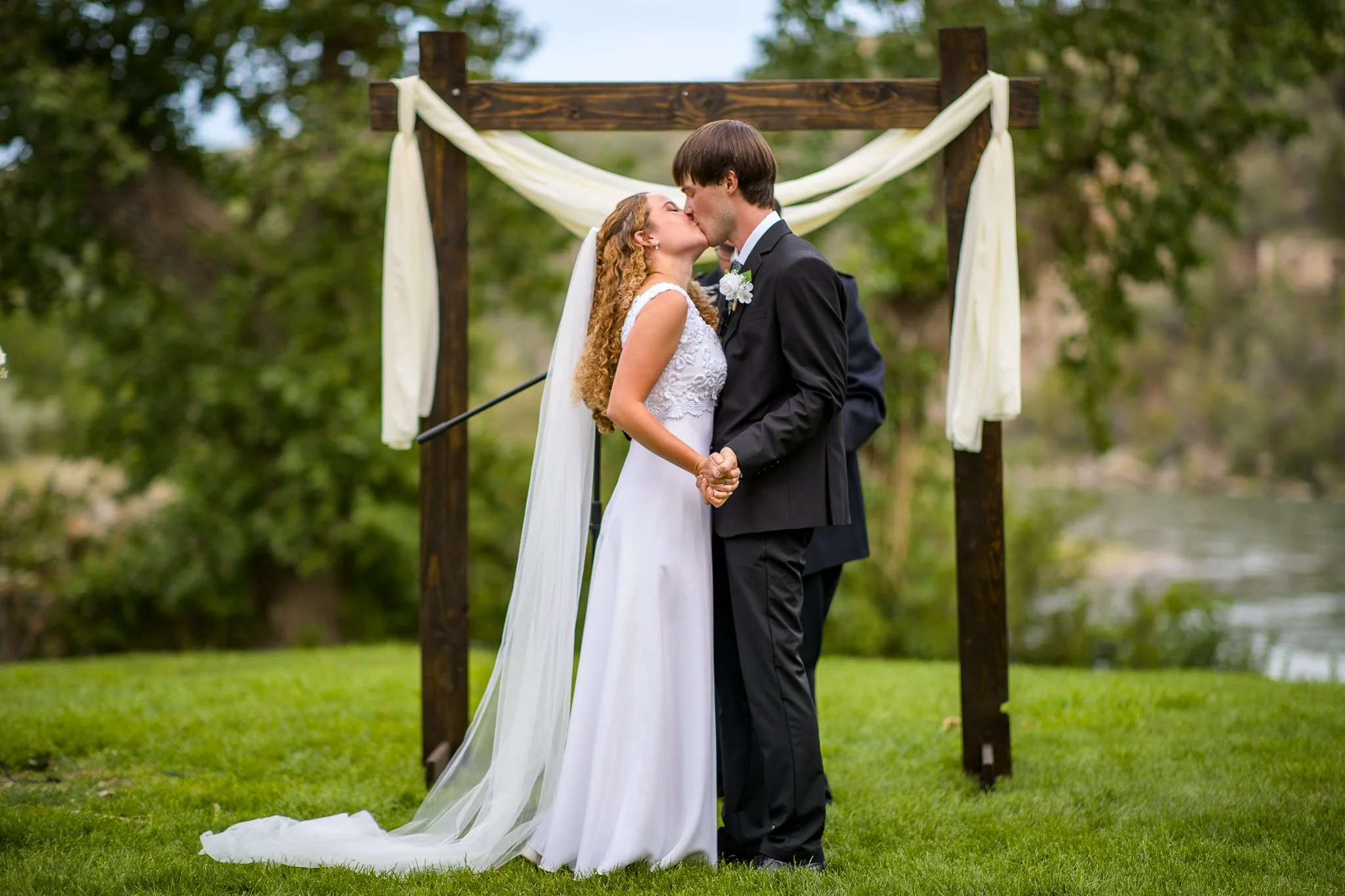 A newlywed couple shares a kiss during their outdoor wedding ceremony, holding hands, with a wooden arch decorated with white fabric in the background, on green grass near a river, surrounded by trees.