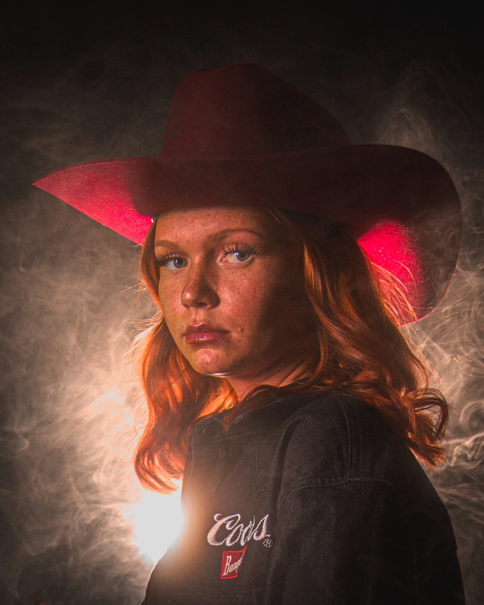 Portrait of a woman with red hair wearing a large cowboy hat and a dark denim jacket with a small embroidered logo.