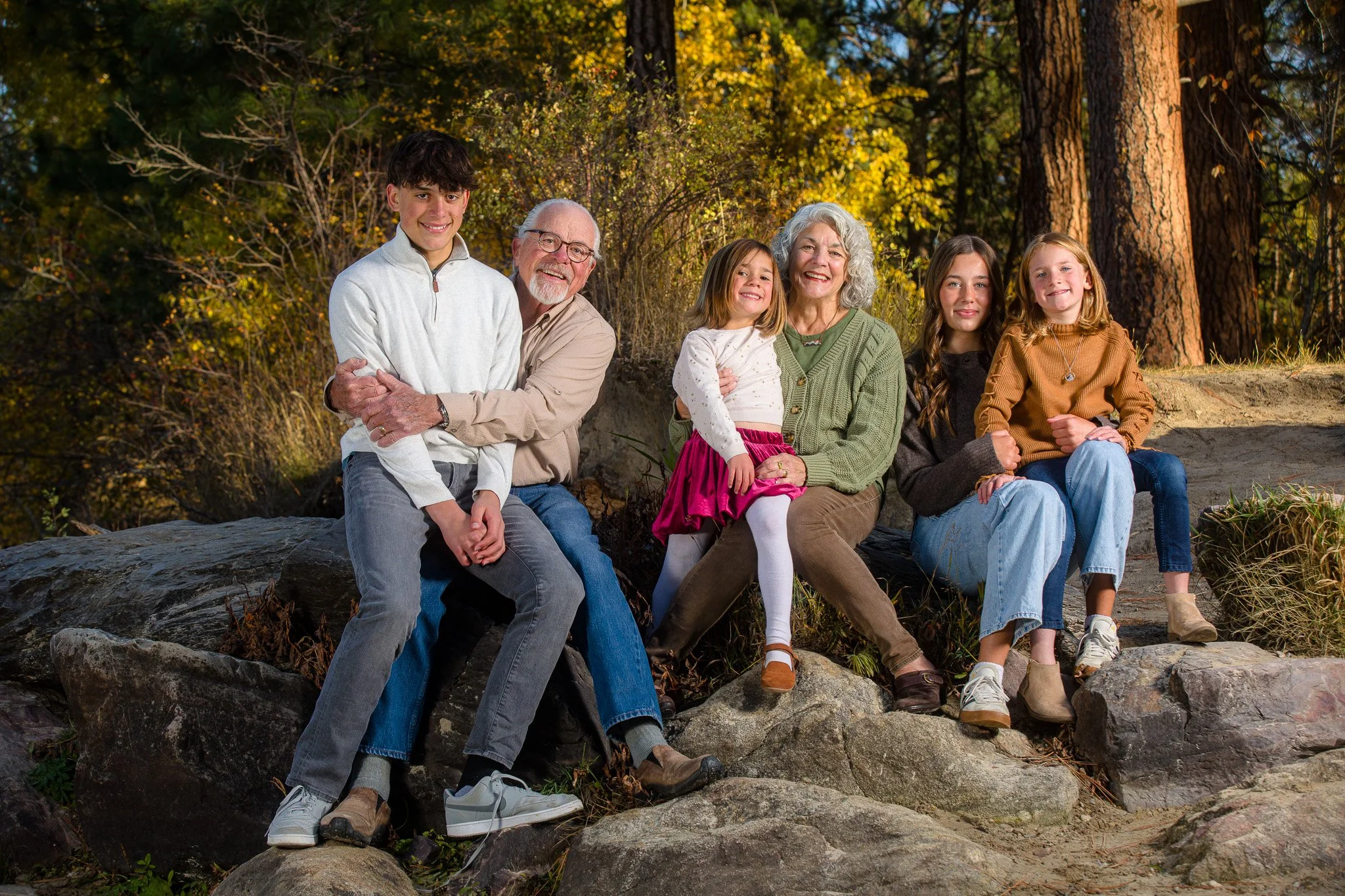 A multigenerational family of six sitting on rocks outdoors during autumn, smiling and enjoying the sunlight.