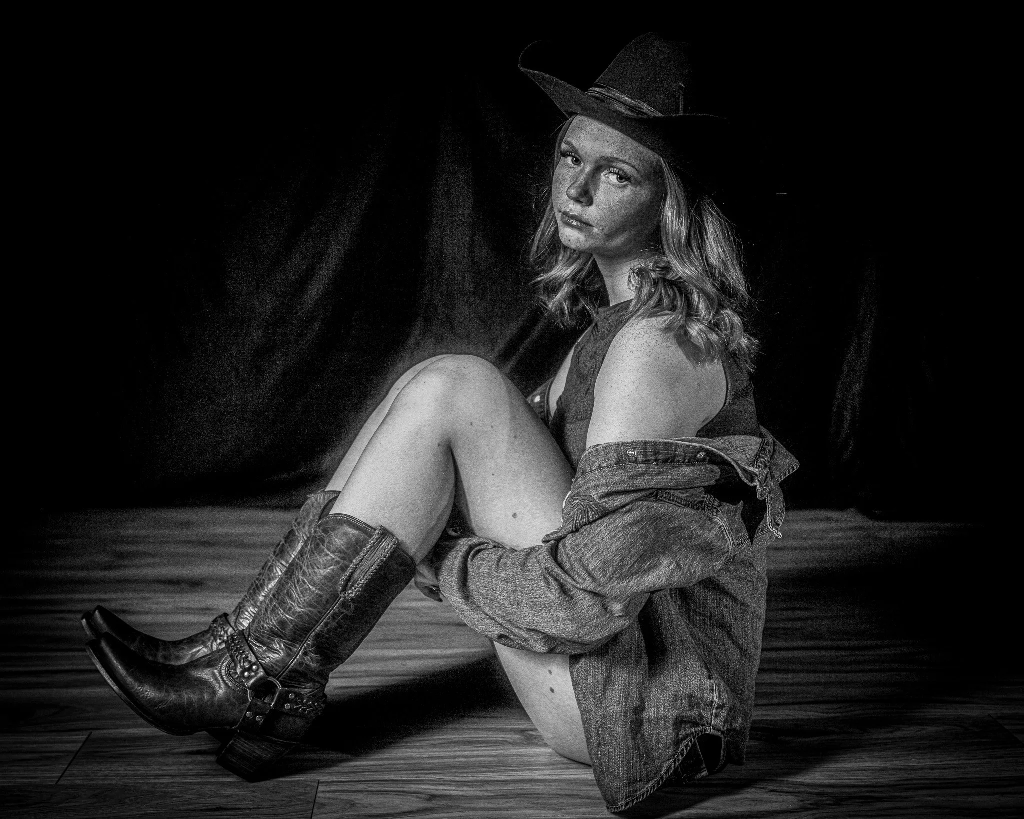 Black and white photo of a woman sitting on wooden floor, wearing a cowboy hat, denim jacket off her shoulders, and cowboy boots, with a dark background.