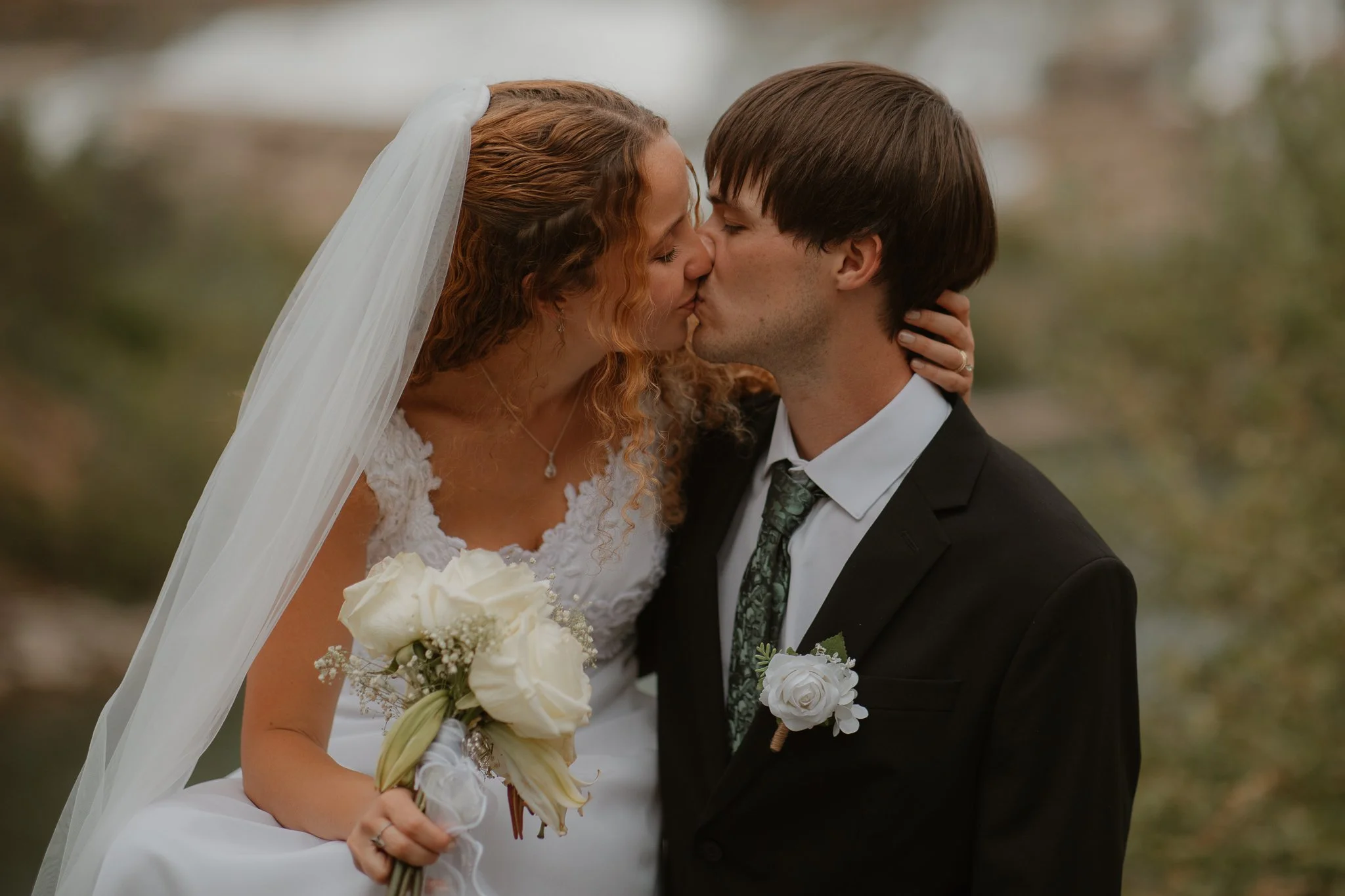 A bride and groom kissing outdoors, the bride holding a bouquet of white roses and wearing a white wedding dress with lace details, and the groom wearing a black suit with a white shirt and a floral tie.