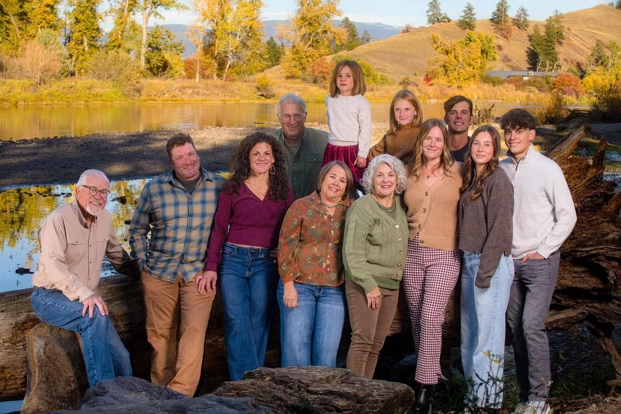 A multigenerational family of fifteen people, including children and seniors, gathered outdoors near a river with colorful fall foliage and rolling hills in the background.
