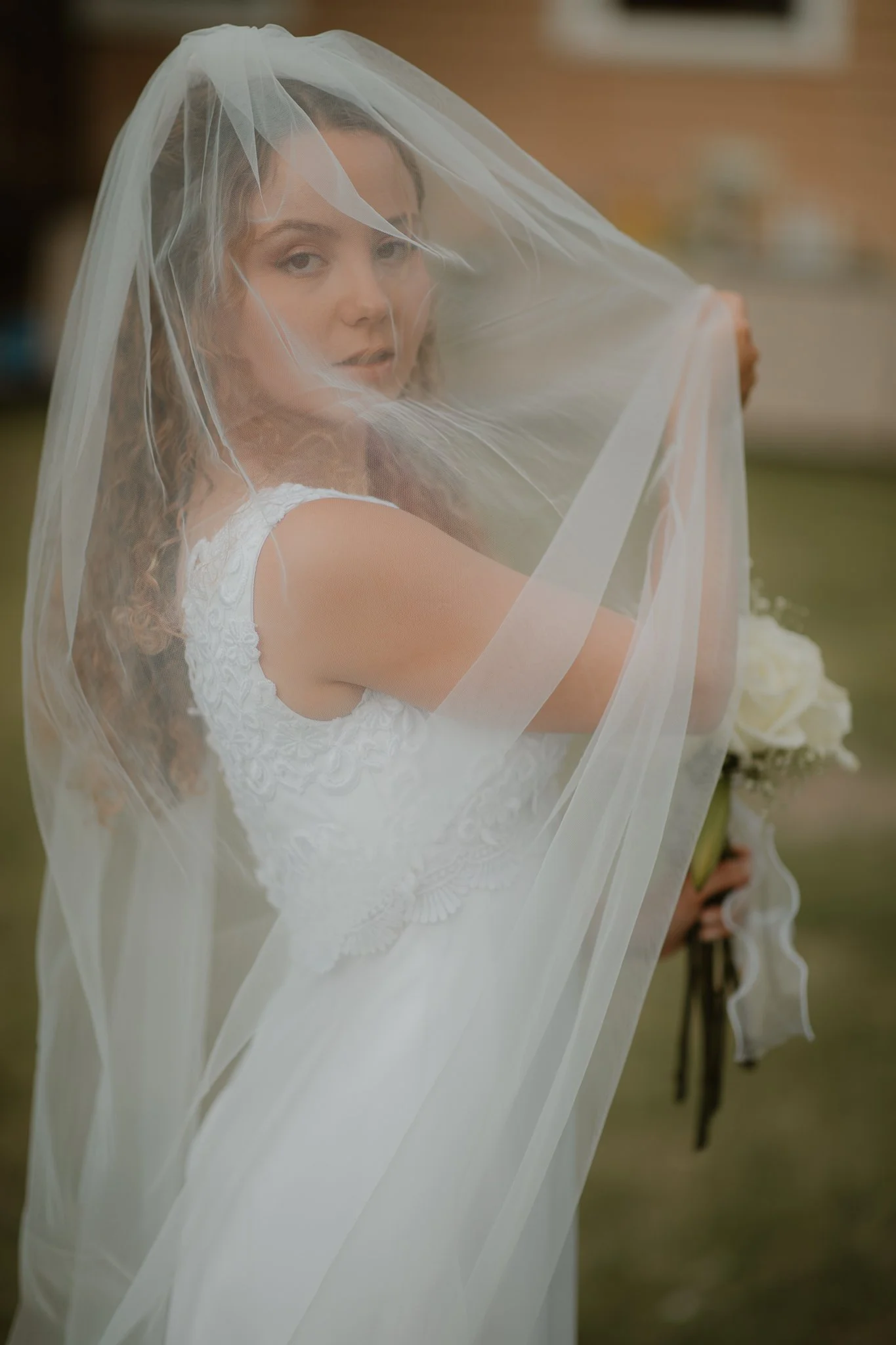 A bride in a white wedding dress and veil holding a bouquet of white flowers, standing outdoors.