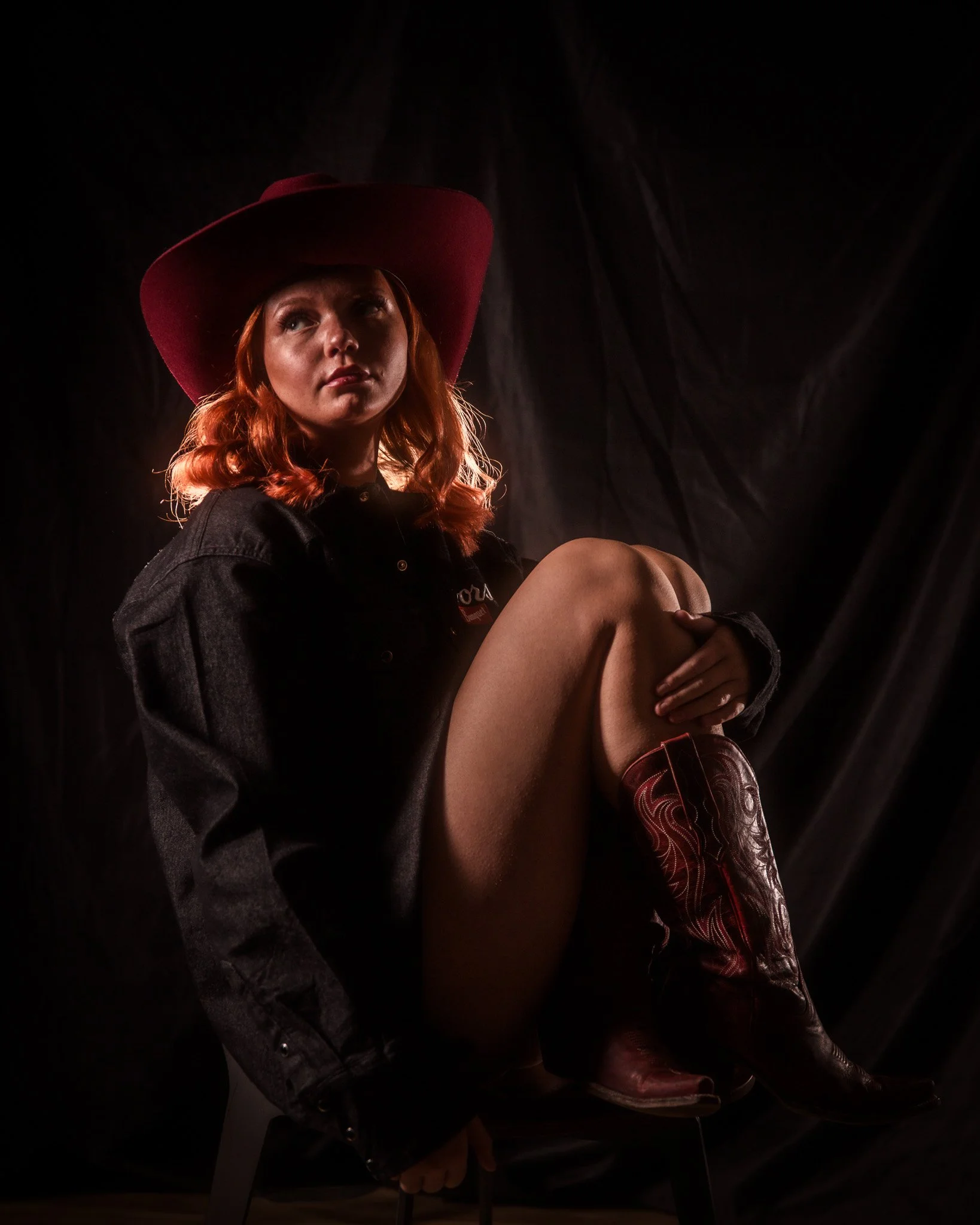A woman with red hair wearing a large red hat, dark jacket, and red cowboy boots. She is sitting on a chair with one leg crossed over the other, posing against a dark background.