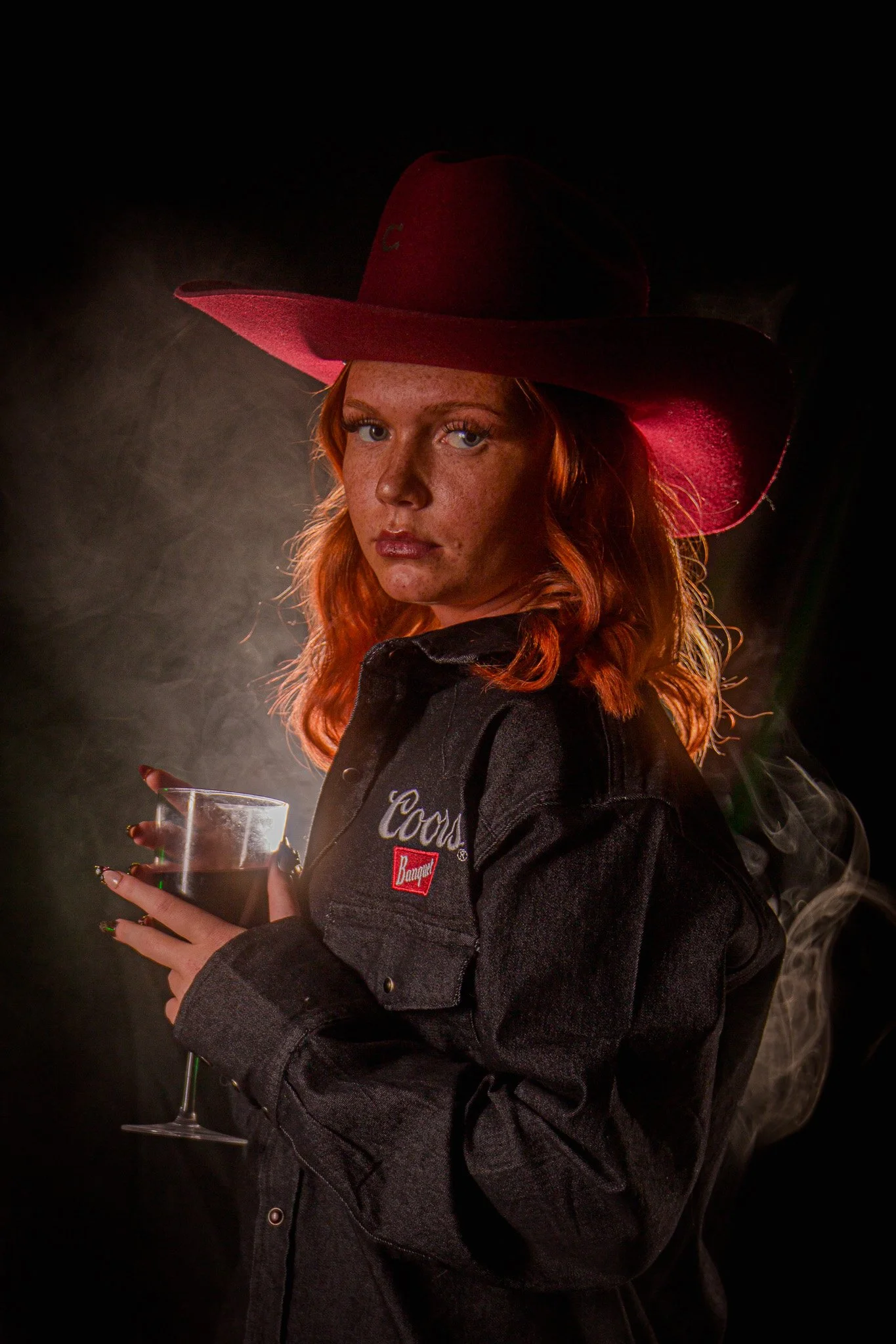 A woman with red hair and freckles wearing a red cowboy hat and a black denim jacket holding a glass of red wine, with smoke in the background.