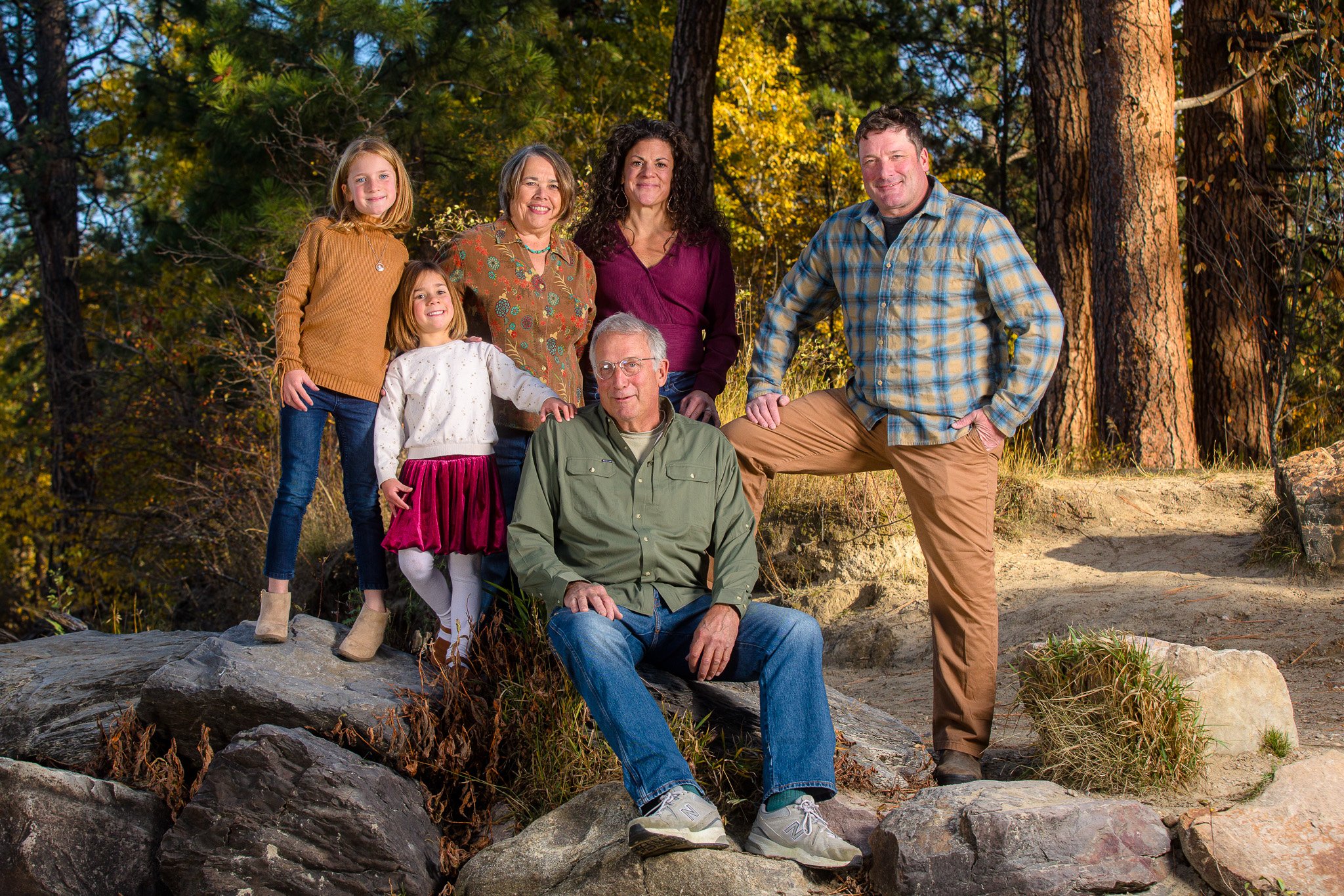 A multigeneration family of six posing outdoors on rocks in a forest with trees and autumn foliage in the background.