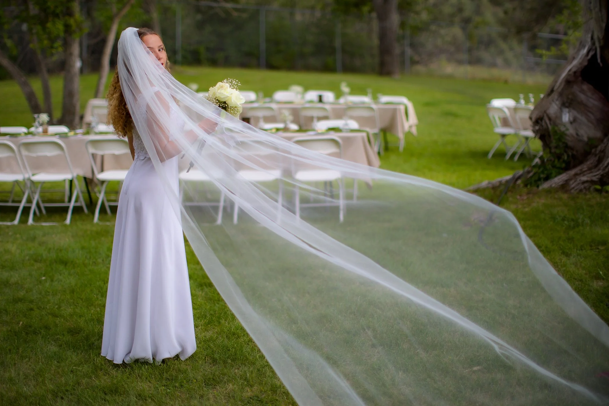 A bride in a white wedding dress and long veil stands on a grassy lawn holding a bouquet of white roses, with empty tables and chairs set up outdoors in the background.
