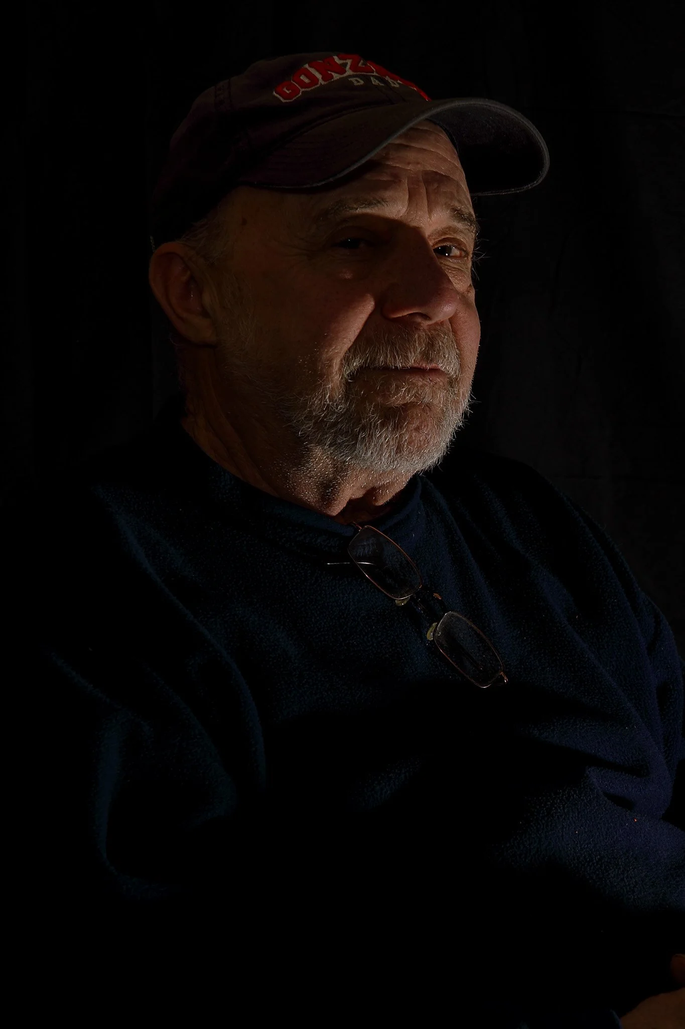 A portrait of an older man with a gray beard and mustache, wearing a baseball cap and glasses, against a dark background.