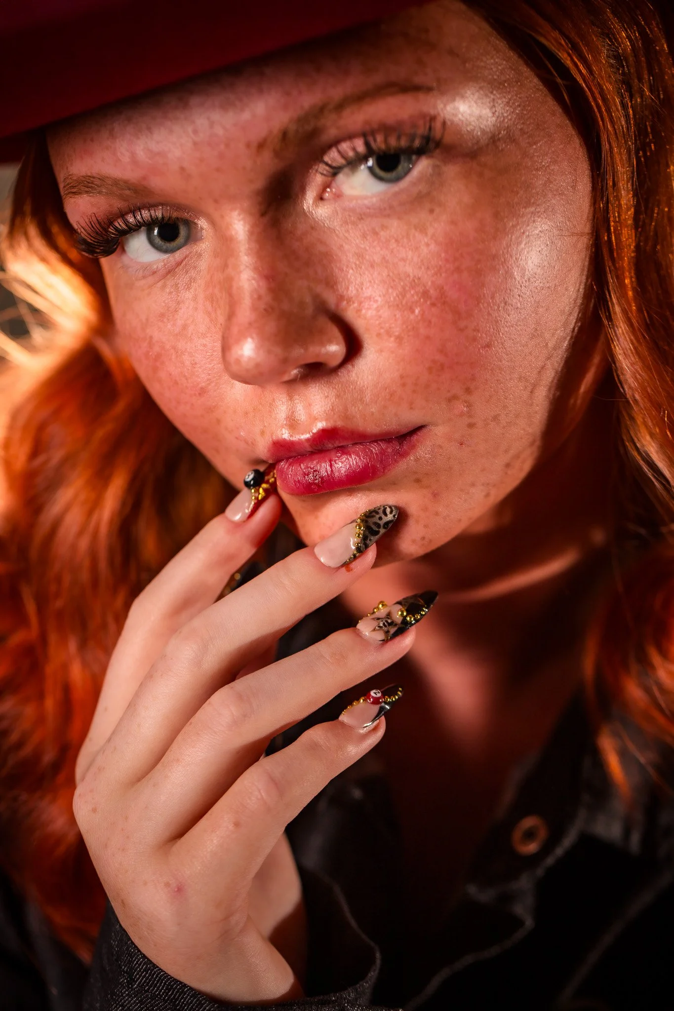 Close-up of a woman with red hair, light freckles, blue eyes, and painted nails, wearing a black hat and jacket.