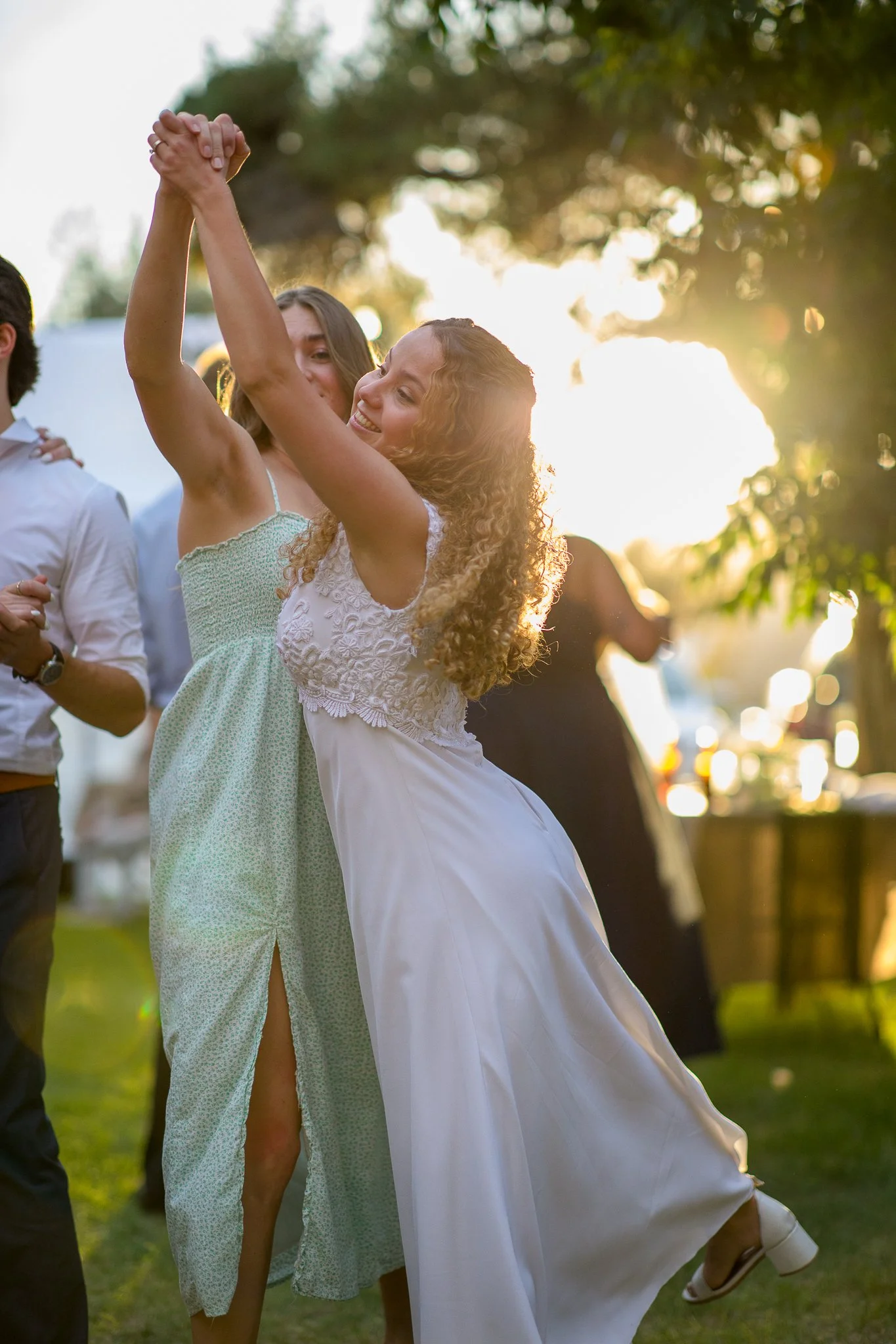 Two women dancing outdoors during sunset, one in a light green dress and the other in a white dress, smiling and holding hands.