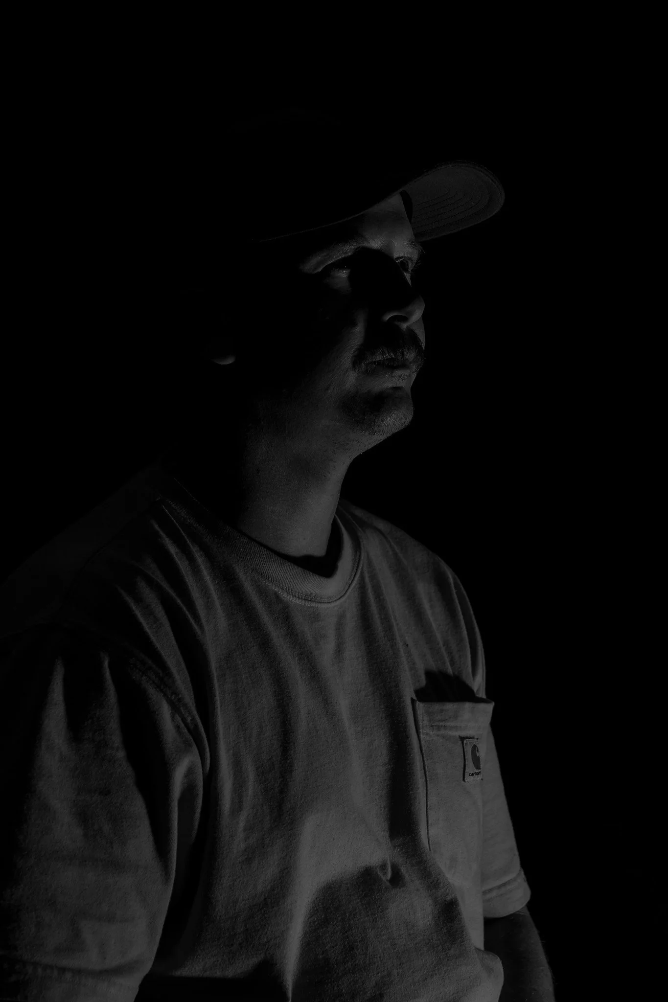 A black and white portrait of a man wearing a baseball cap and a t-shirt, with his face partially illuminated against a dark background.
