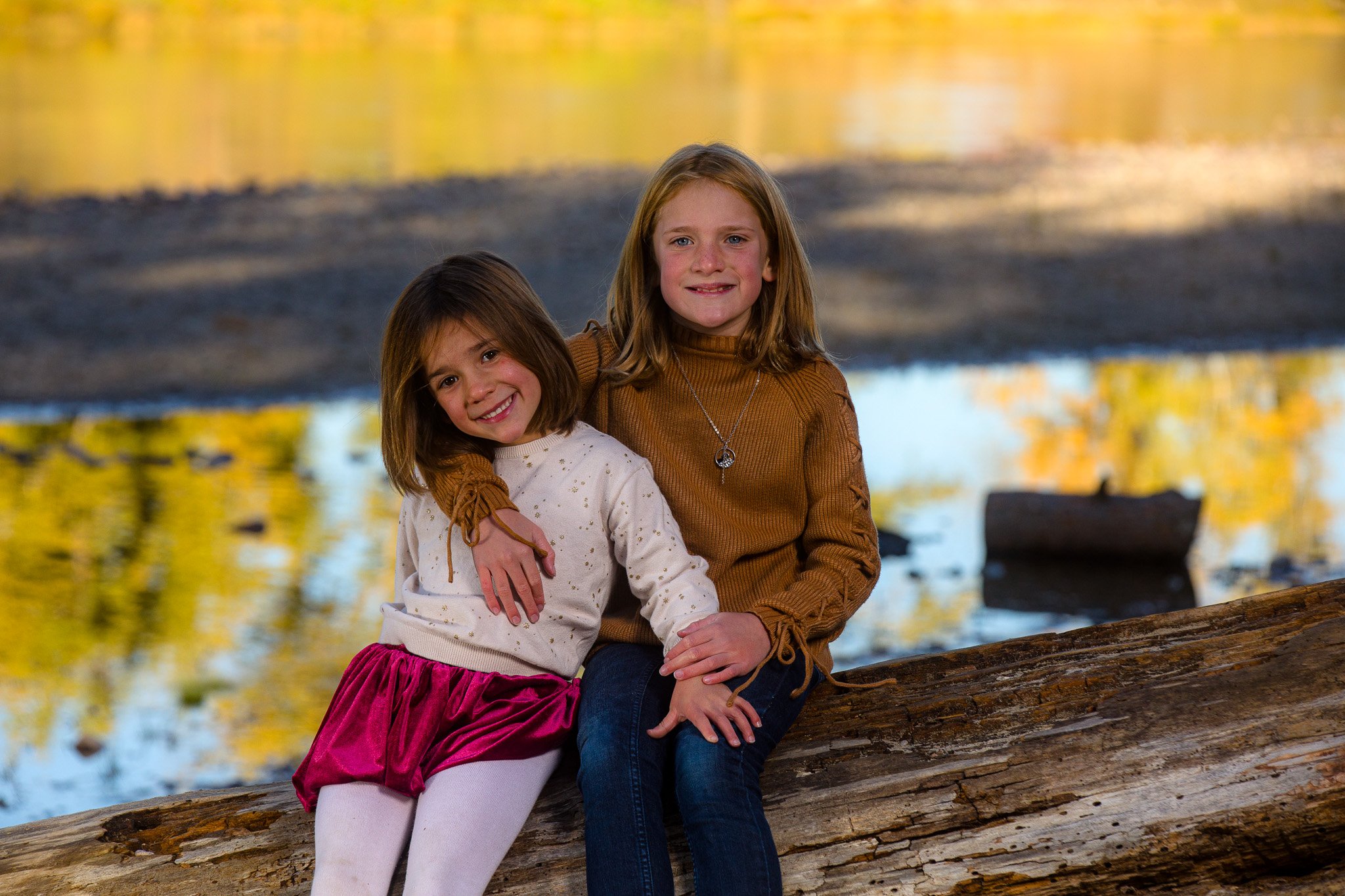 Two young girls sitting on a large fallen log beside a body of water in a forested area with autumn foliage in the background. One girl is smiling and wearing a white sweater and a pink skirt, the other girl is smiling and wearing a brown sweater and