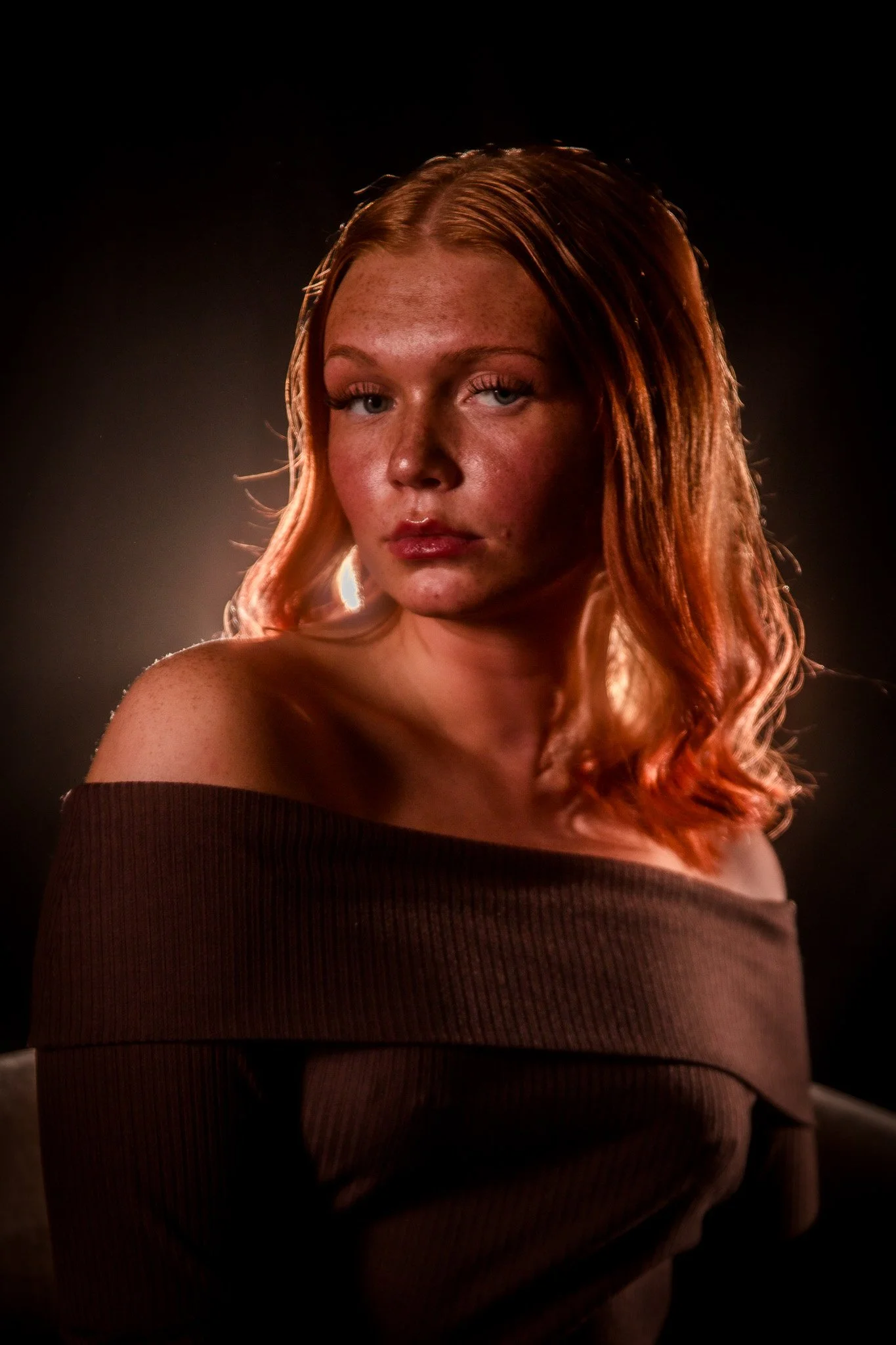 Portrait of a young woman with long, wavy red hair, wearing an off-the-shoulder brown top, against a dark background with subtle lighting.