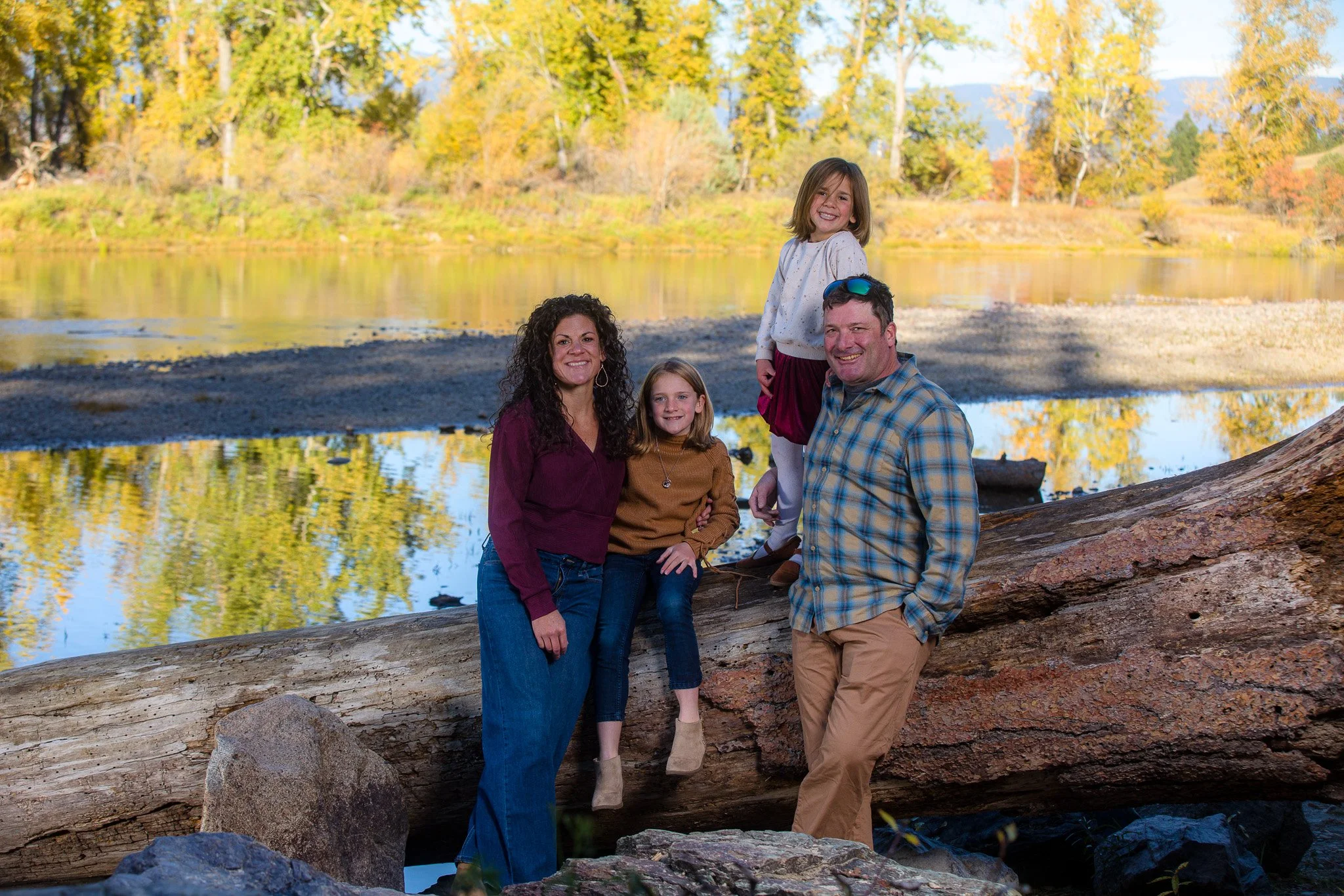 A family of four, including two women and two girls, posing on and around a fallen log near a river with autumn trees in the background.