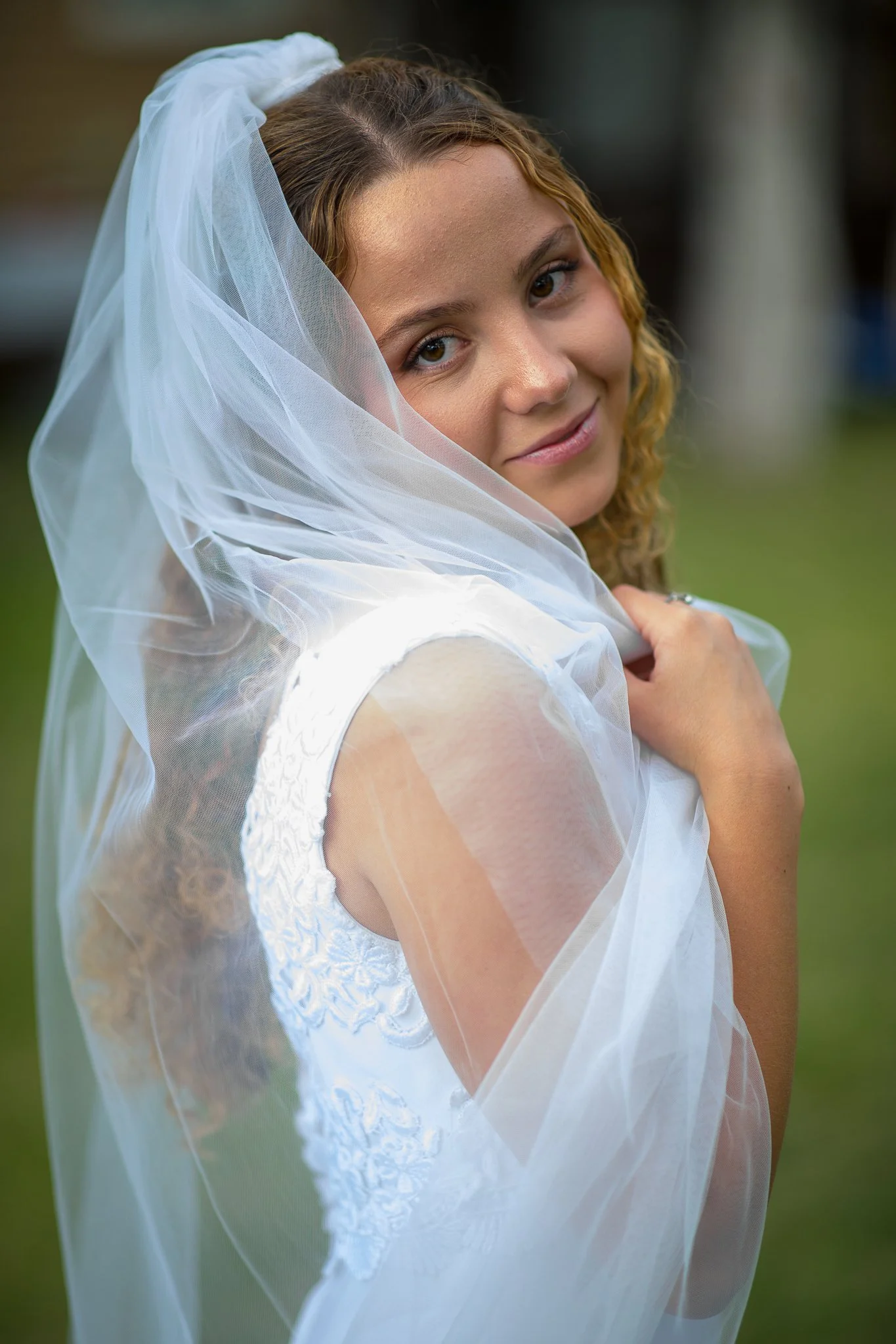 A young woman with curly hair is wearing a wedding dress and veil, looking at the camera with a gentle smile, outdoors on a cloudy day.