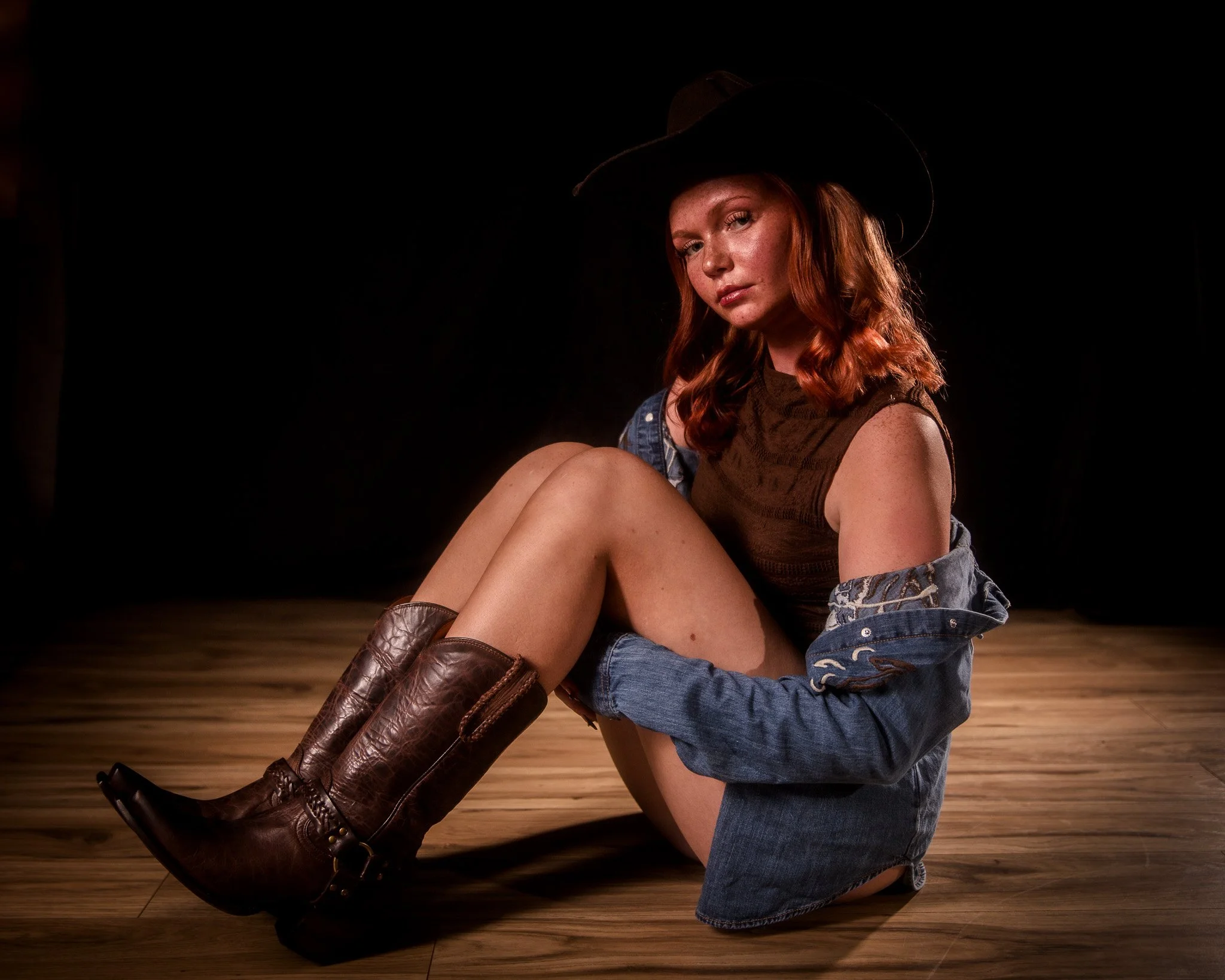 Young woman with red hair wearing a black cowboy hat, brown sleeveless top, denim jacket, and brown cowboy boots, sitting on a wooden floor against a black background.