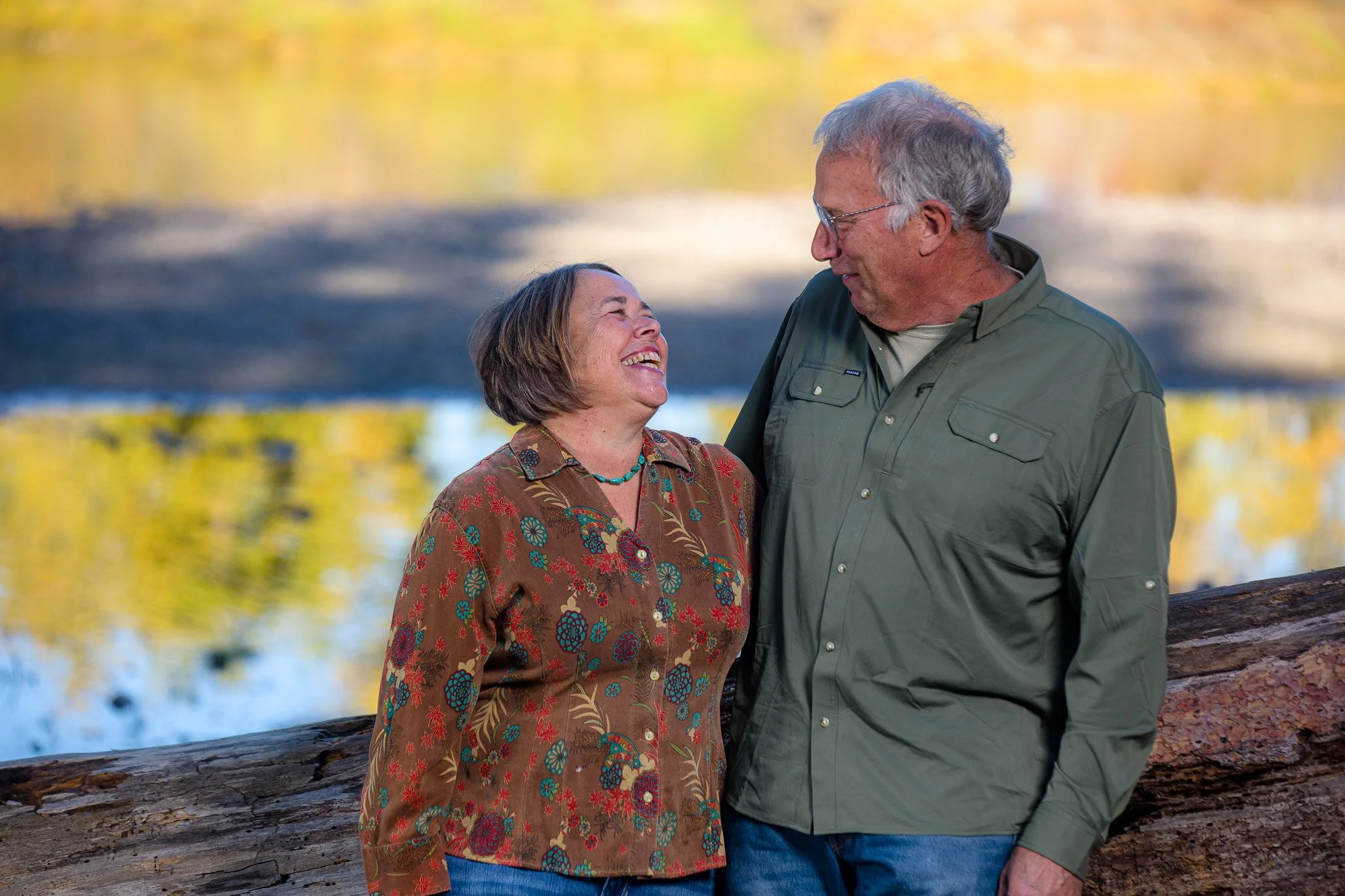A smiling older woman and man standing outdoors near a log, looking at each other, with a blurred autumn-colored background and a body of water.