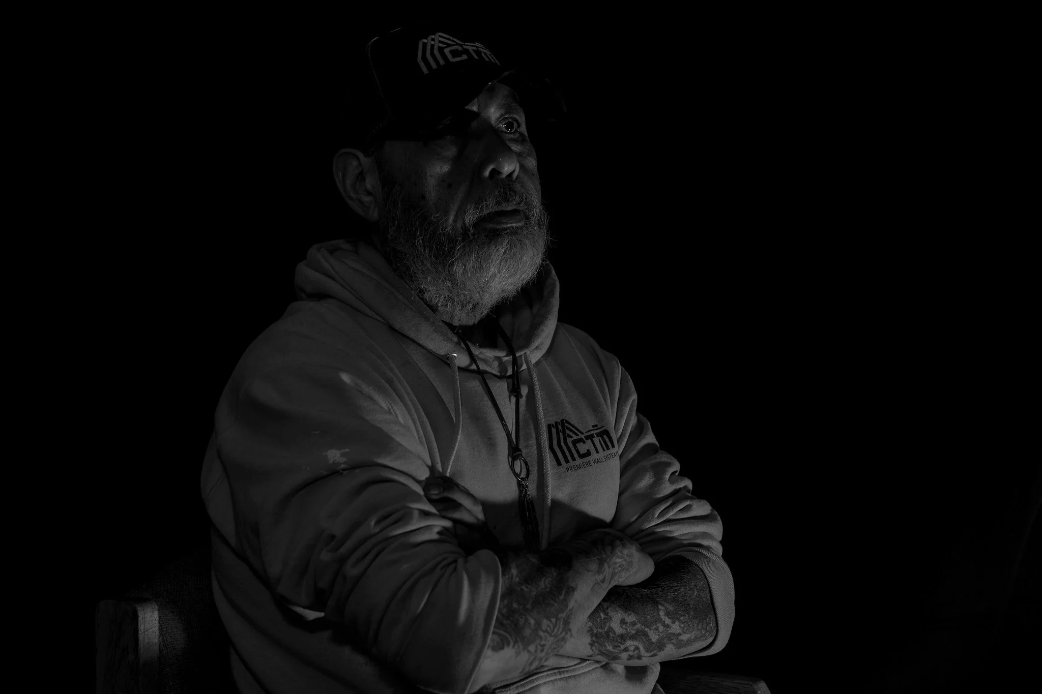 A black-and-white photo of an older man with a beard, wearing a hoodie and a cap, sitting with his arms crossed against a dark background.