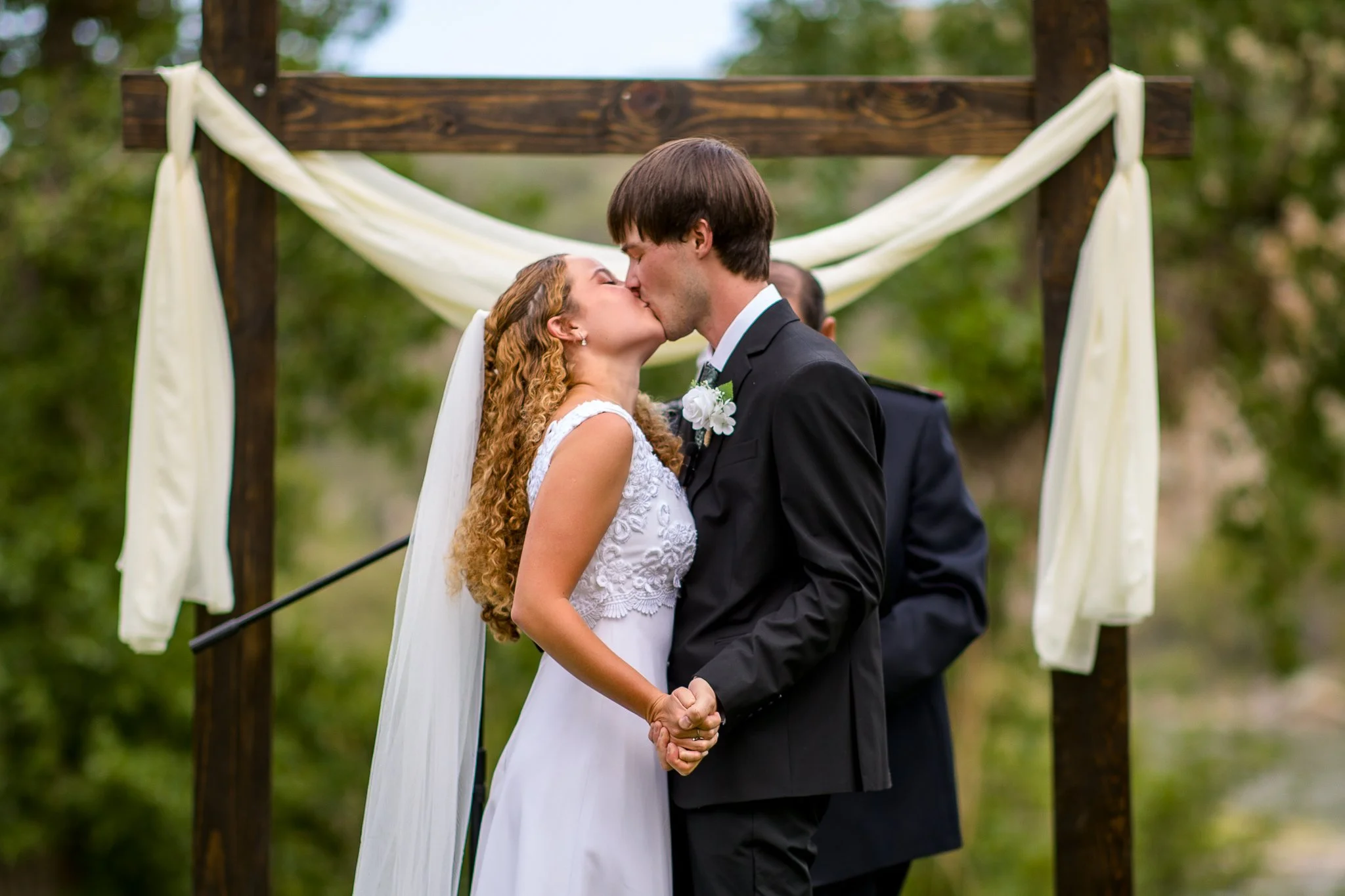 A bride and groom kissing during their outdoor wedding ceremony, holding hands, with a wooden arch decorated with light-colored fabric in the background.