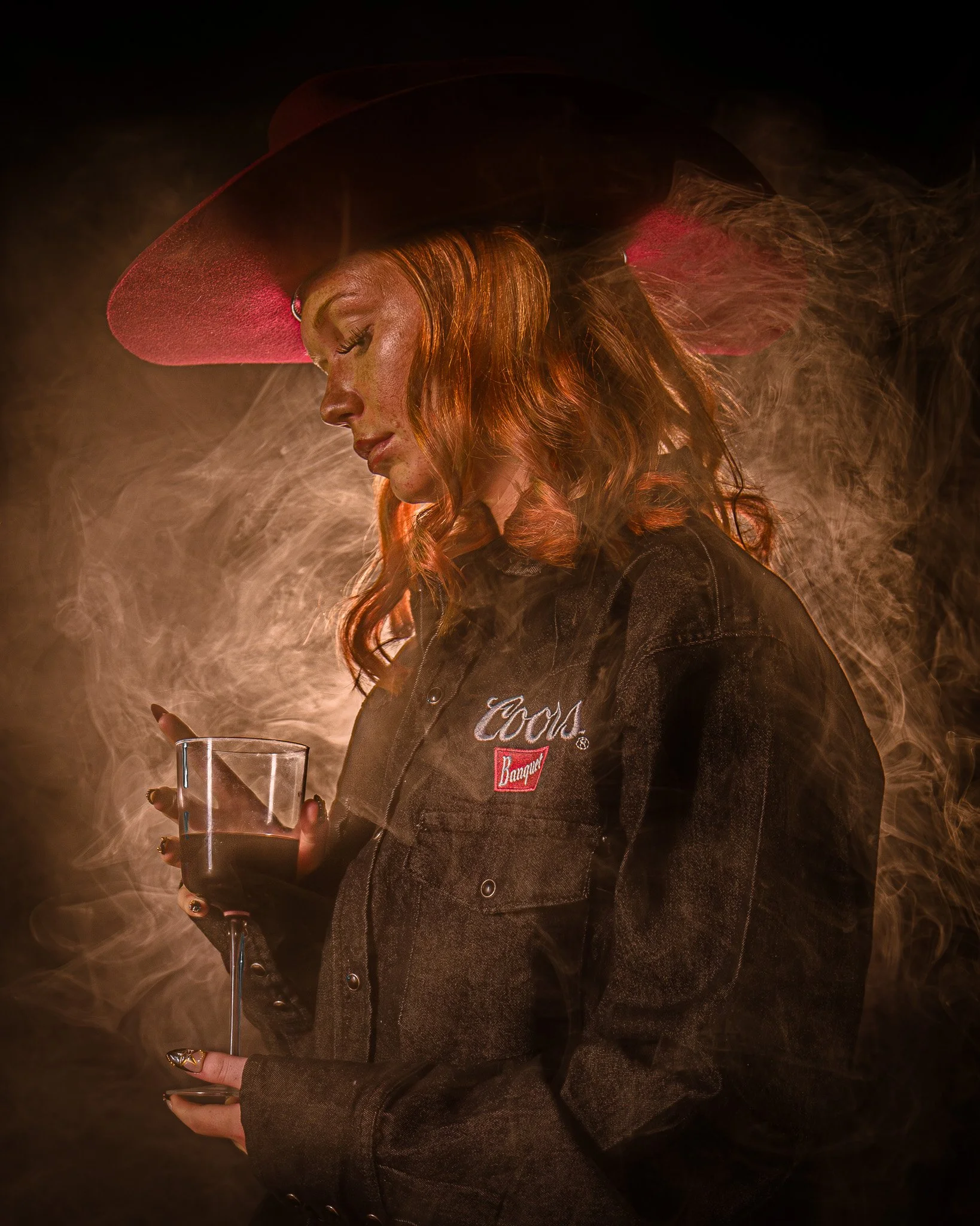 A woman with red hair wearing a wide-brimmed hat and a dark jacket holding a glass of red wine, with smoke surrounding her.