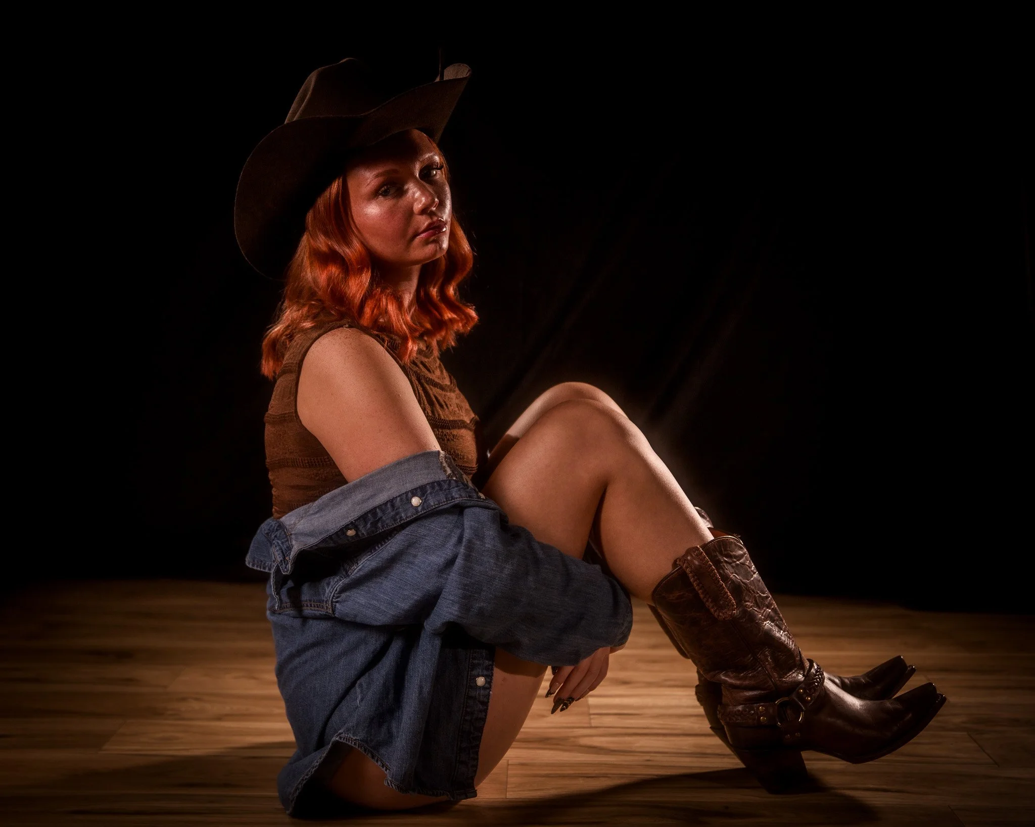 Woman with red hair wearing a cowboy hat, sleeveless top, partially covered denim jacket, and cowboy boots, sitting on a wooden floor against a black background.