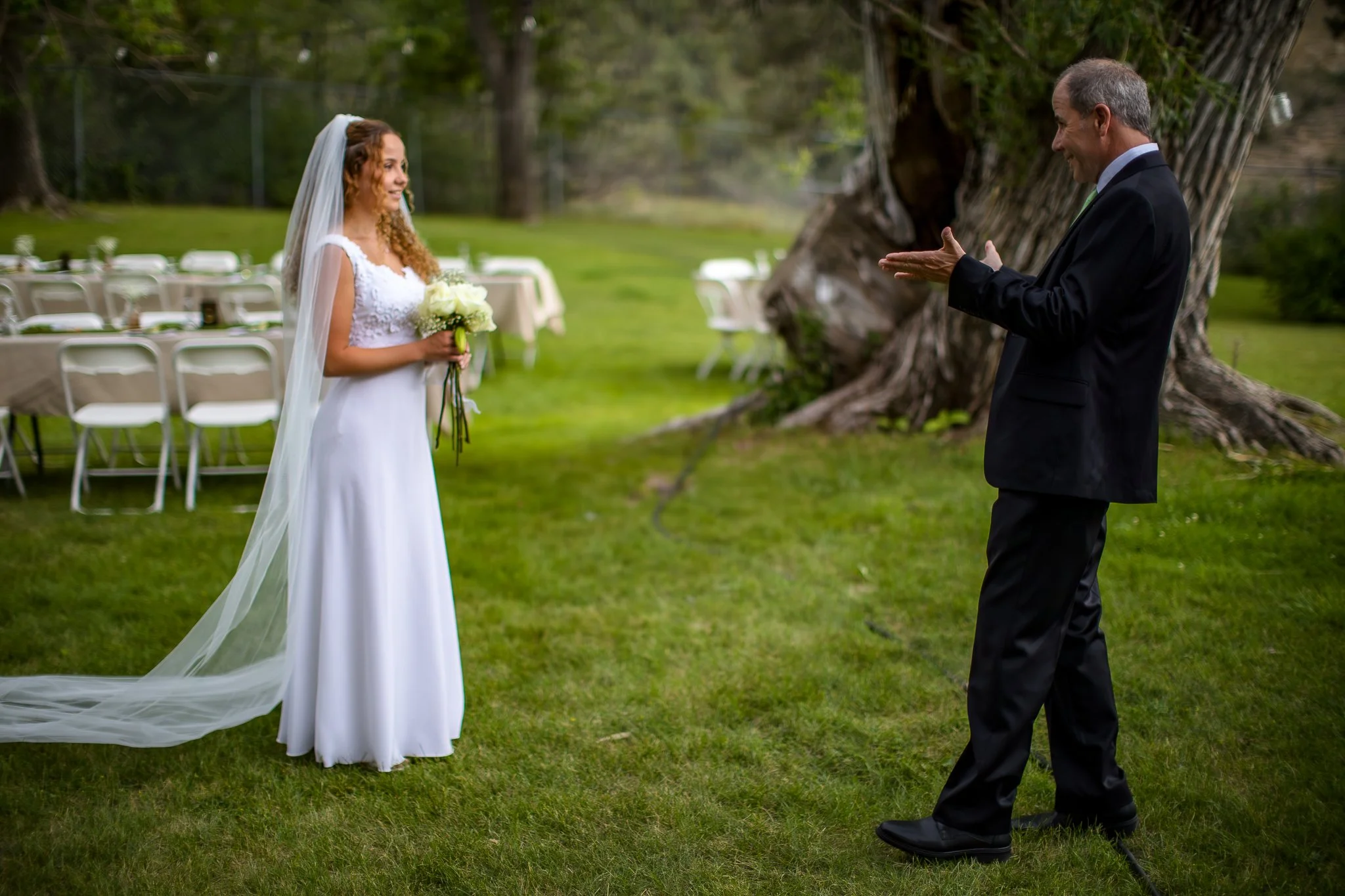 A bride in a white wedding dress with a veil holding a bouquet of white roses standing in front of a man in a black suit, outdoors on a grassy area with tables and chairs in the background.