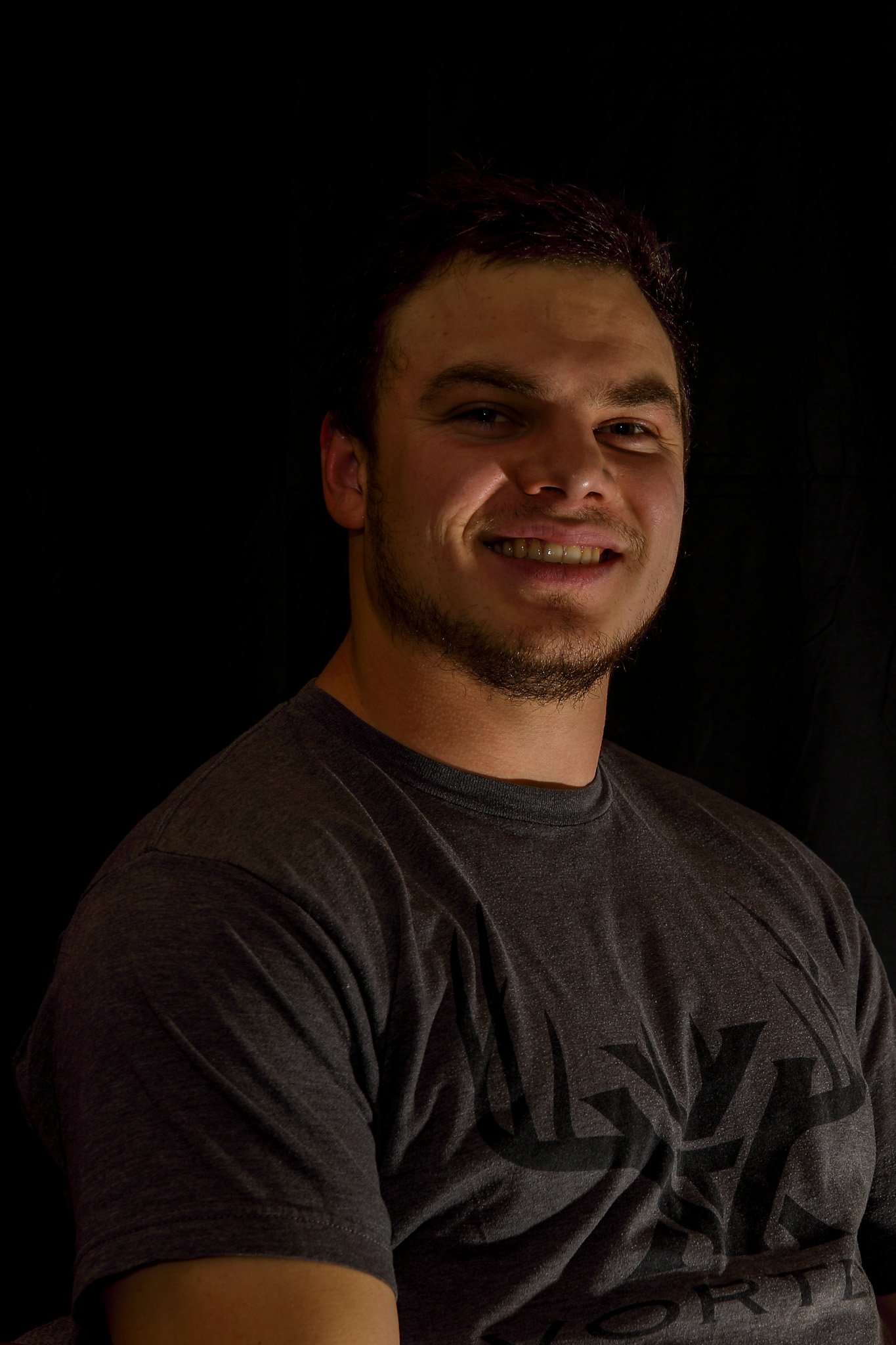 A young man with short dark hair, a beard, and a slight smile, sitting against a black background, wearing a dark T-shirt with a graphic design.