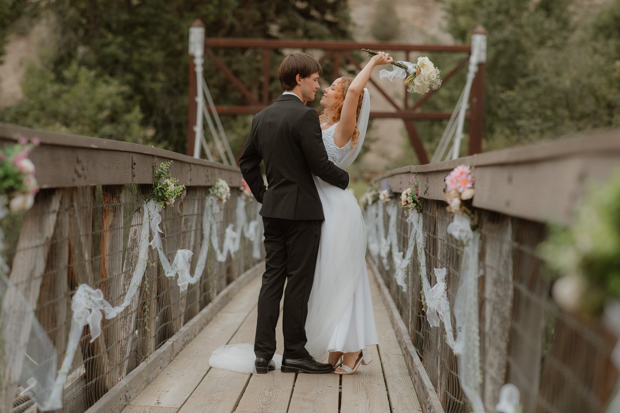 A bride and groom standing on a decorated wooden bridge during their outdoor wedding ceremony, with the bride holding a bouquet and the groom looking at her.