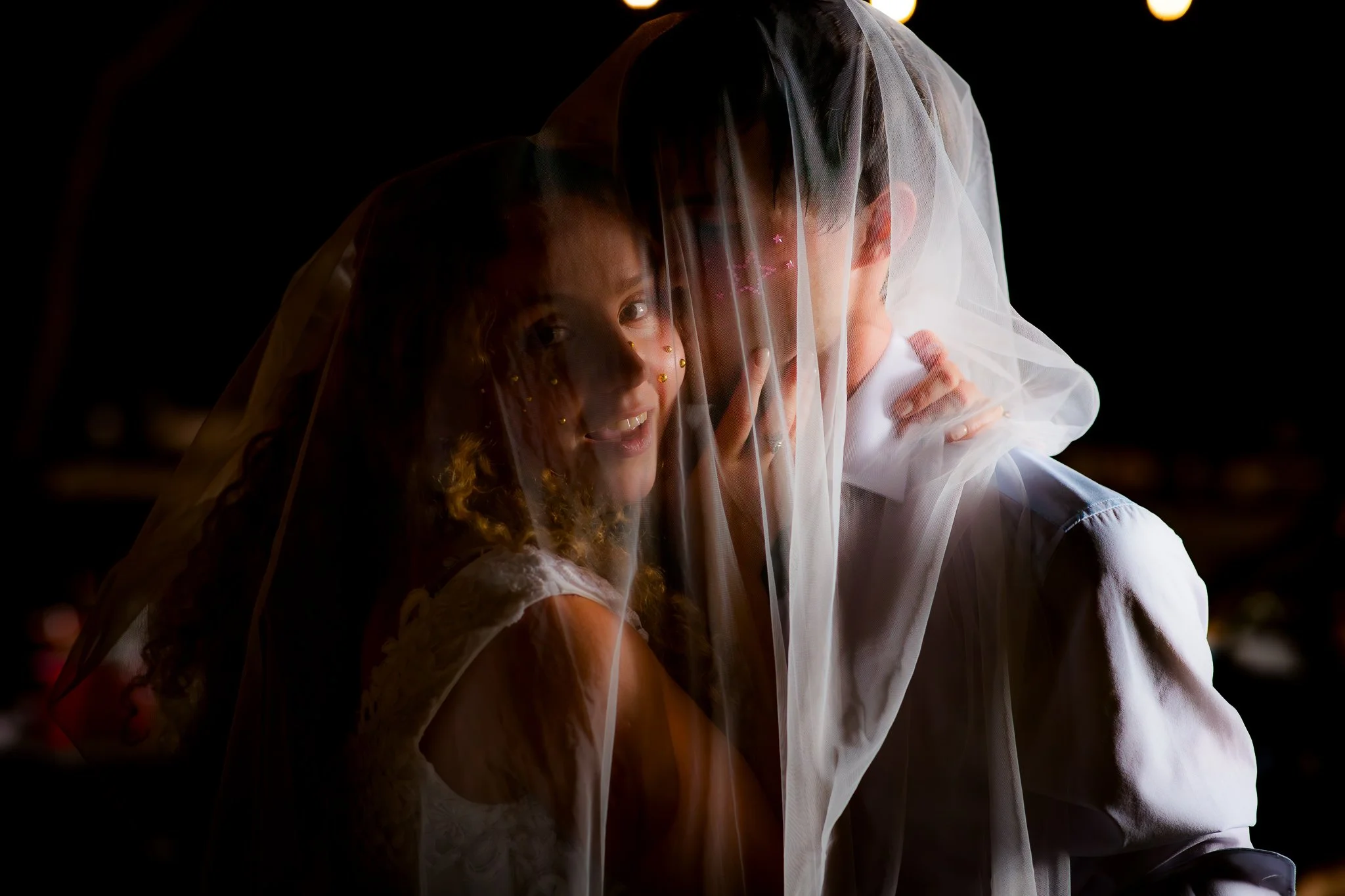 A bride and groom behind a sheer veil, with the bride smiling and the groom partially obscured.