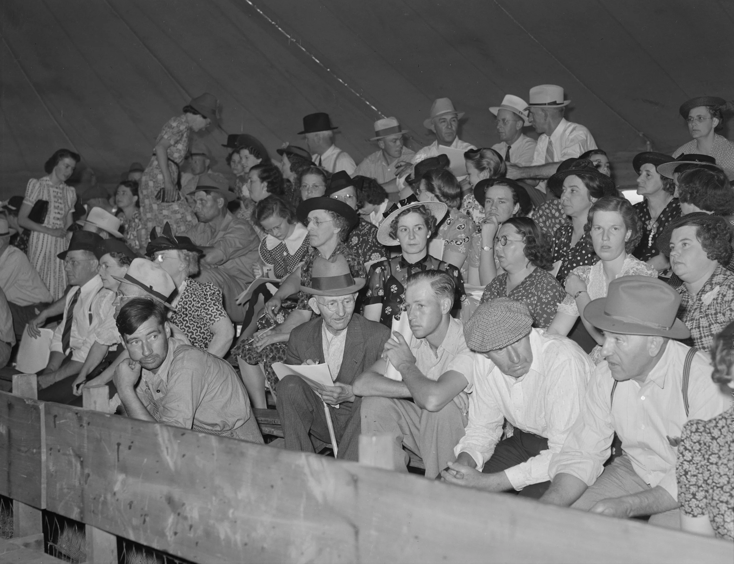 Black and white photo of a crowd of people sitting in a wooden bleacher, many wearing hats and period-style clothing, at what appears to be an outdoor event or game.
