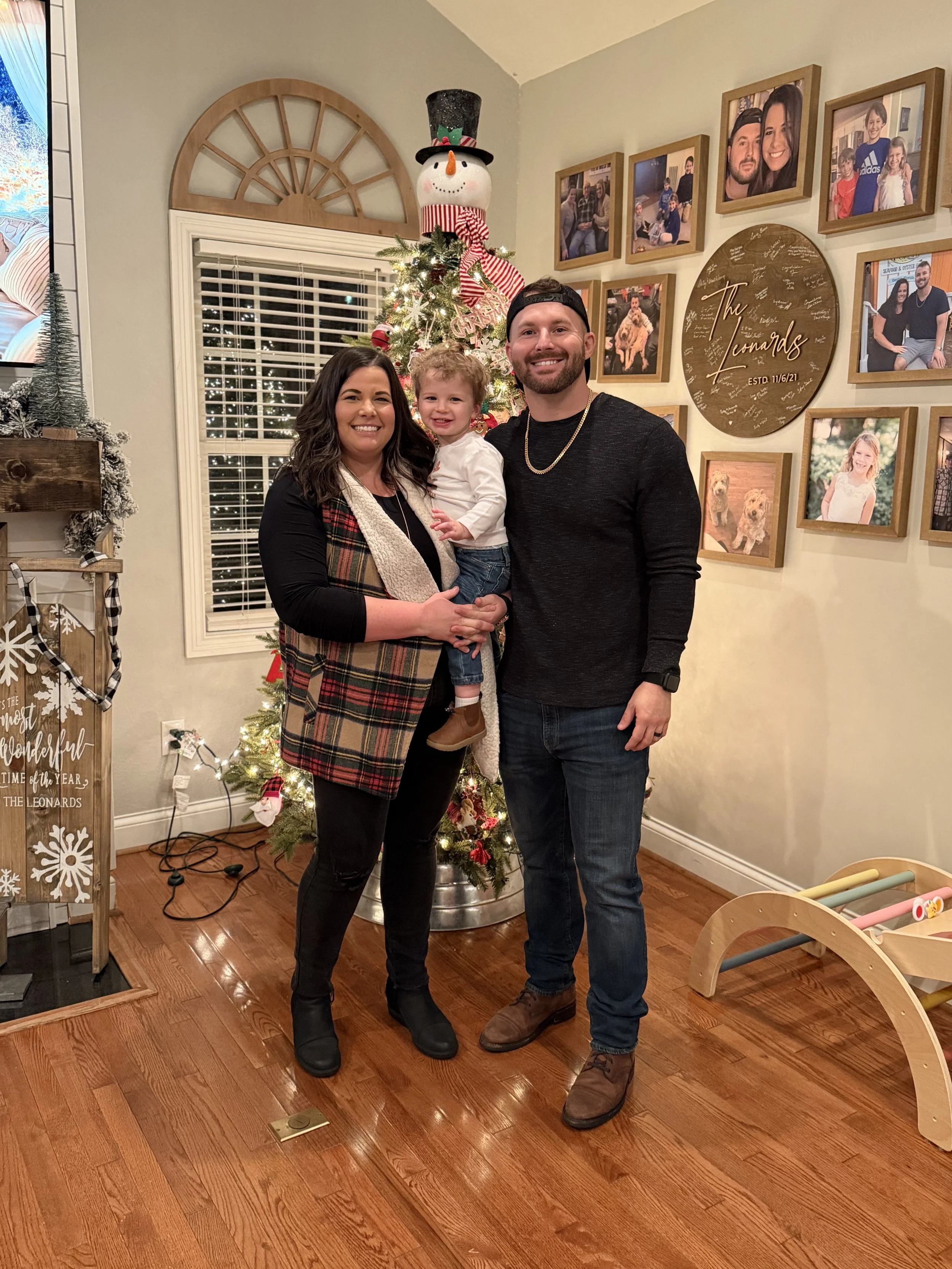A family of three posing in front of a decorated Christmas tree indoors. The woman and man are standing with a young girl, smiling at the camera. The woman has dark hair, wearing a black top, black pants, black boots, and a plaid vest. The man has a beard, wearing a black shirt, blue jeans, and brown shoes. The girl has curly blond hair, wearing a white top, jeans, and brown shoes.