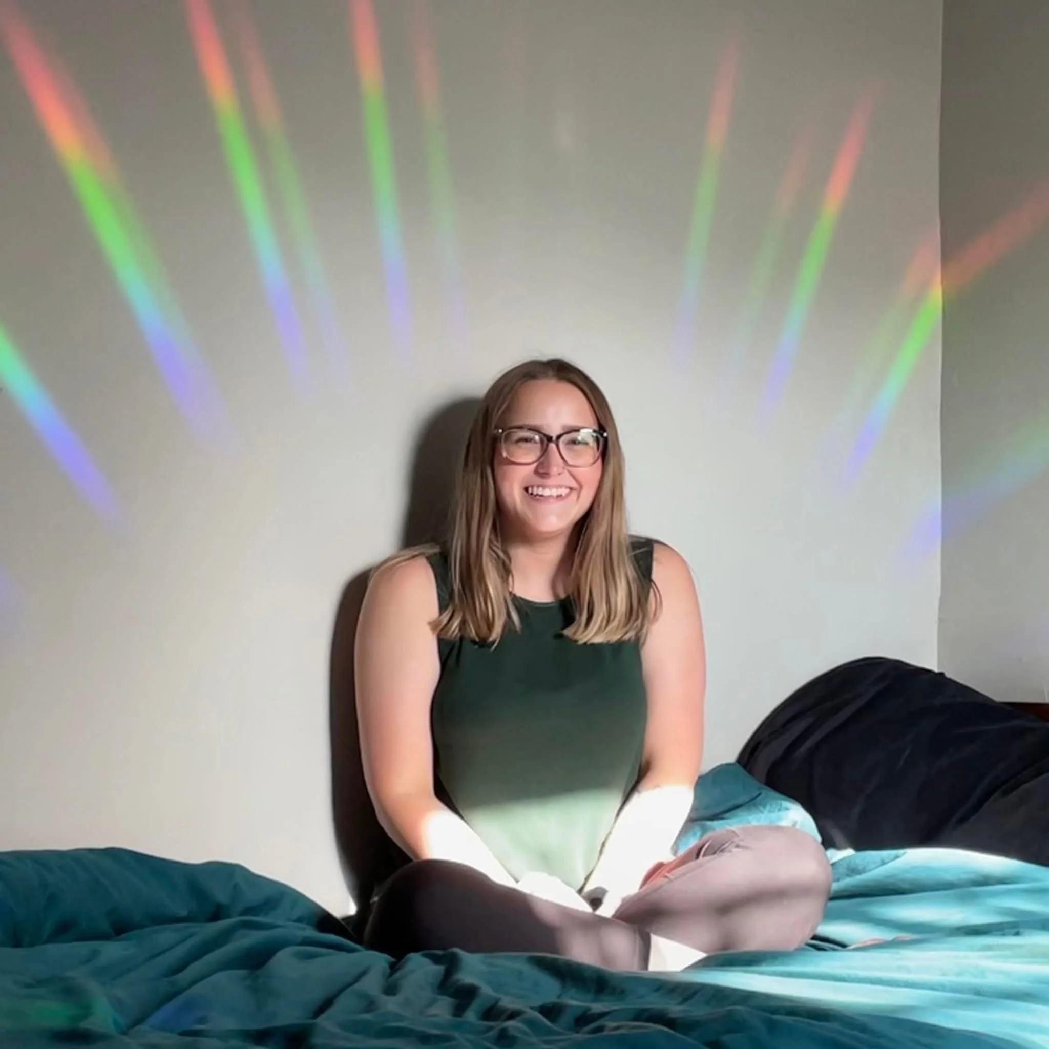 Smiling woman wearing glasses sits cross-legged on a bed beneath colorful rainbow light reflections.