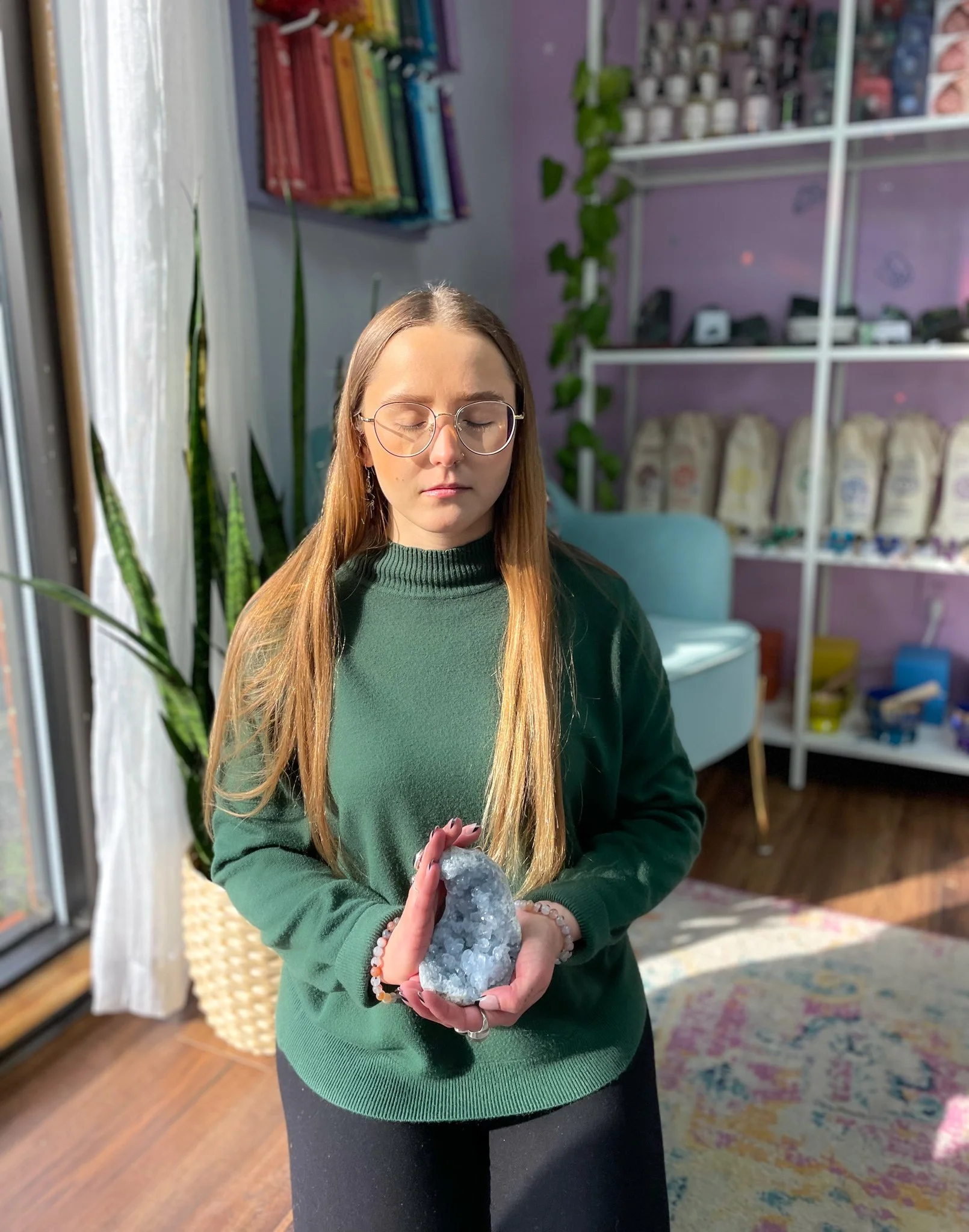 Woman wearing glasses and a green sweater holds a blue crystal cluster in a sunlit room.