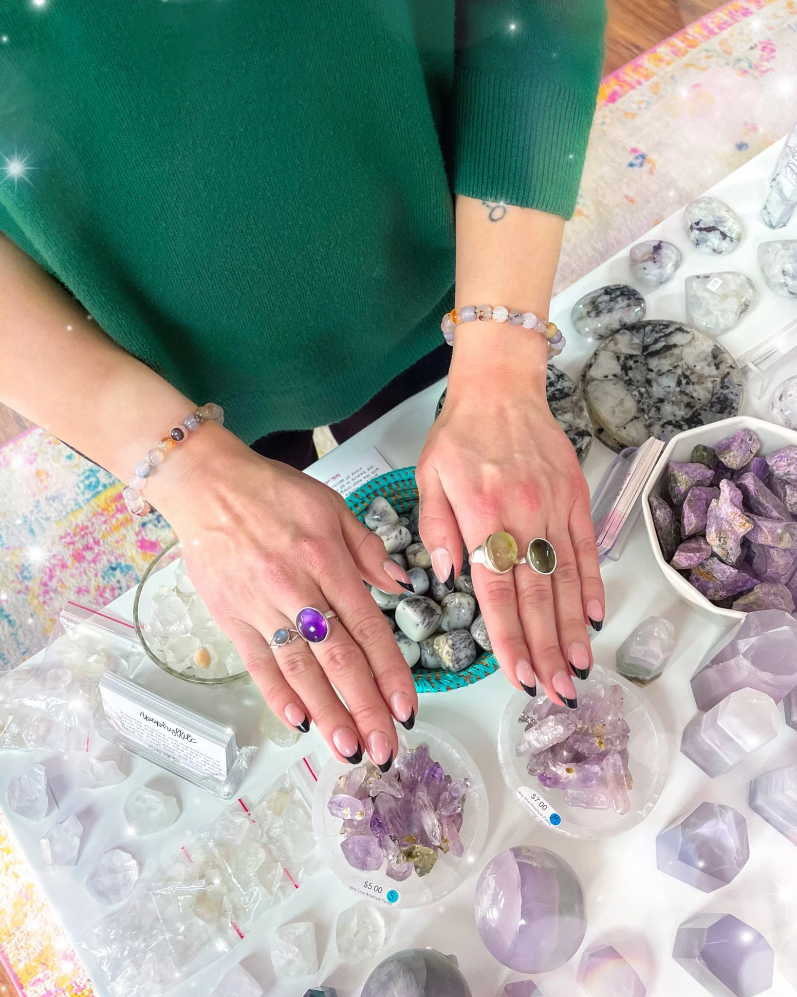 Person wearing gemstone bracelets and rings over a display of polished stones, crystals, and amethyst specimens.