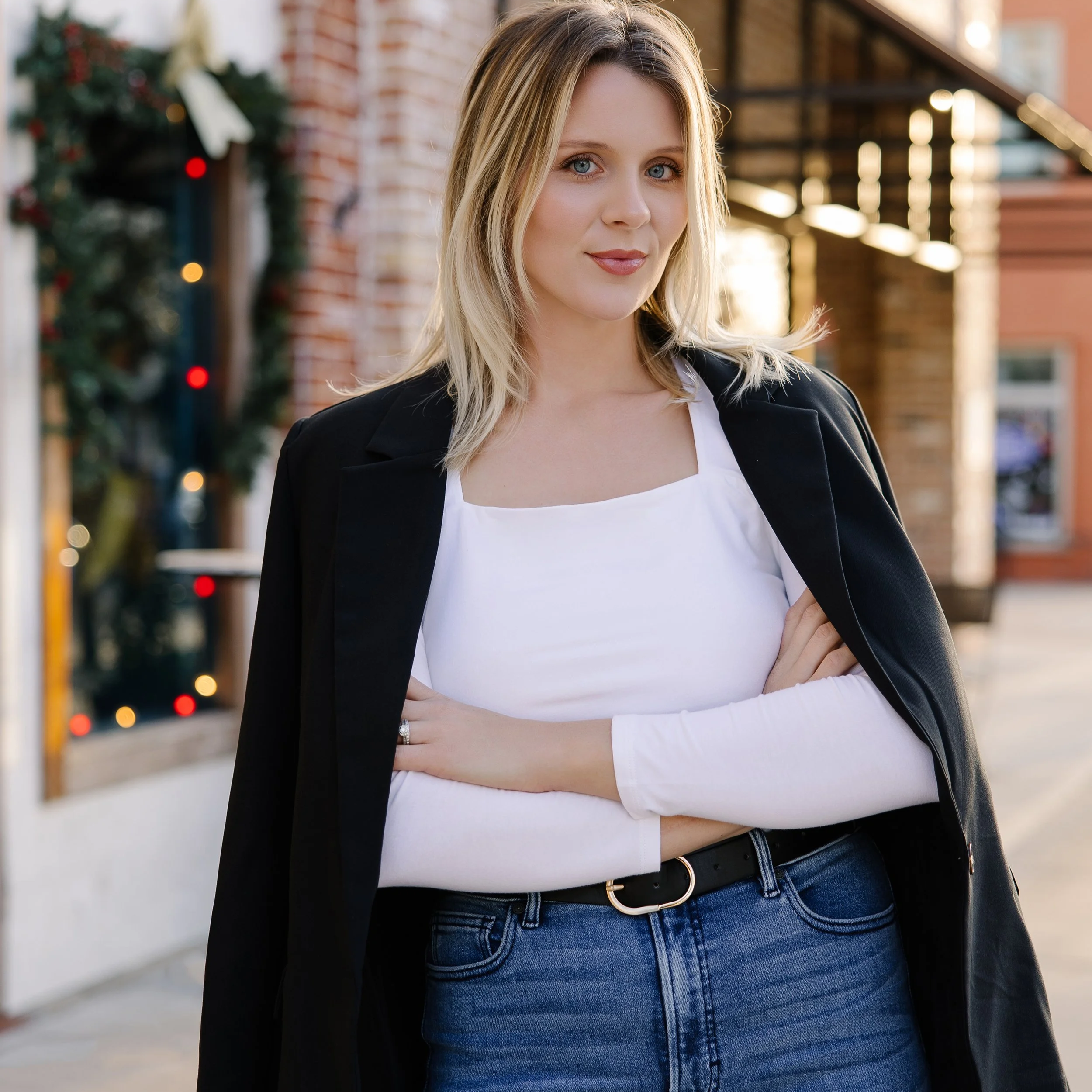 A woman with blonde hair, blue eyes, and fair skin standing outdoors with her arms crossed, wearing a black blazer draped over her shoulders, a white top, and blue jeans, with a brick building and holiday decorations in the background.