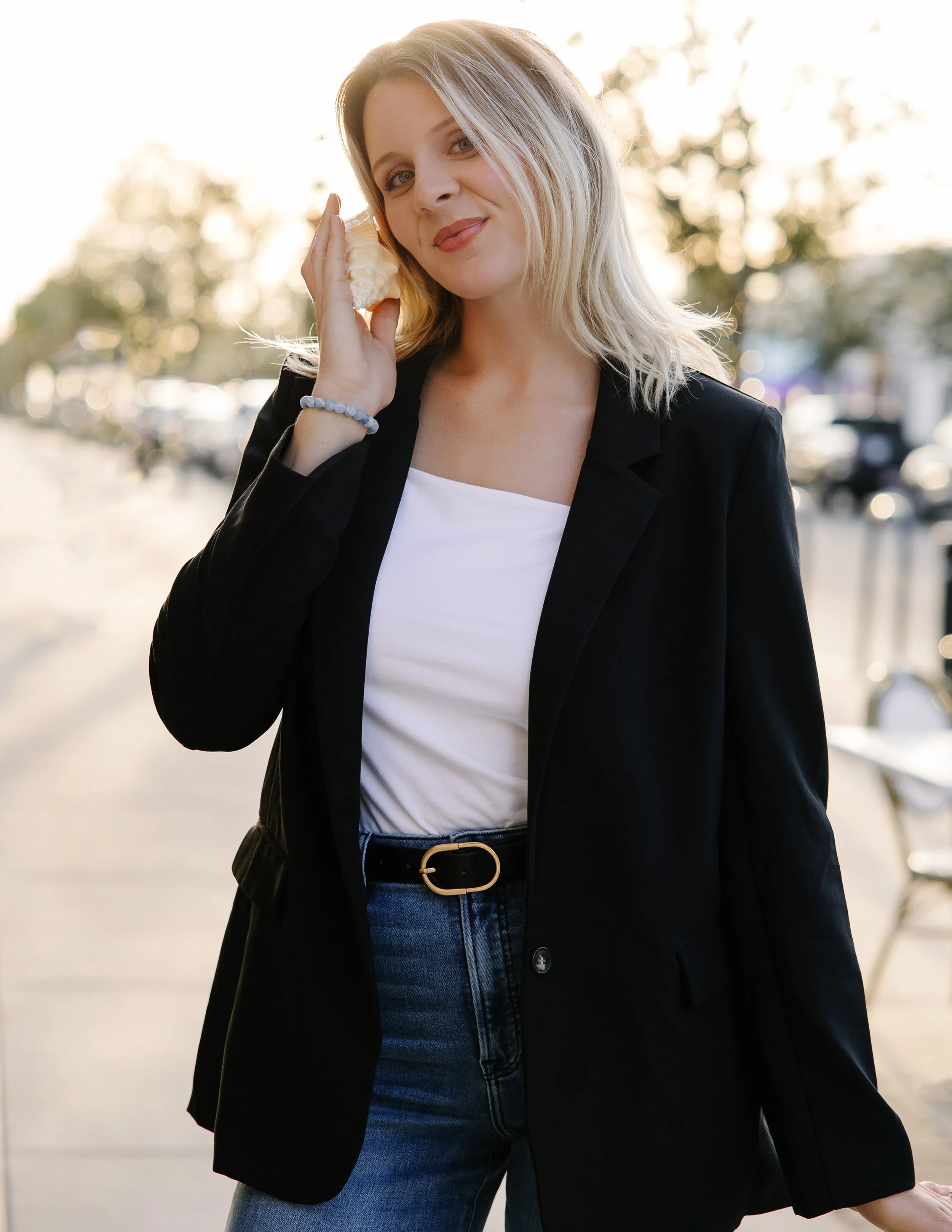 Young woman with blonde hair in a black blazer and jeans, holding a seashell to her ear outdoors during sunset for real estate branding session