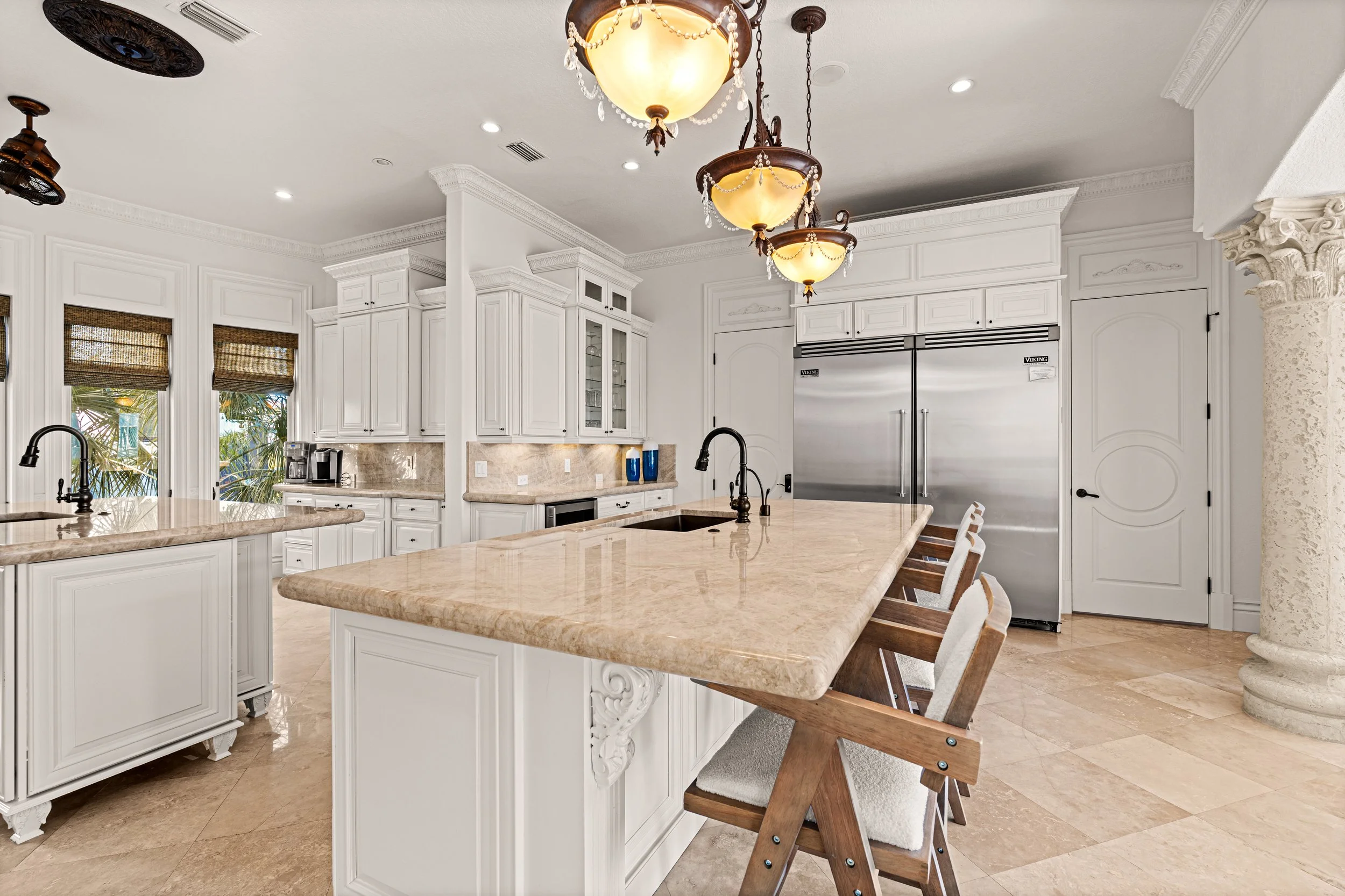 A spacious, elegant kitchen featuring white cabinetry, a large beige marble island, stainless steel double-door refrigerator, and a chandelier with multiple hanging lights. The room has beige tiled floors, crown molding, and windows with woven shades