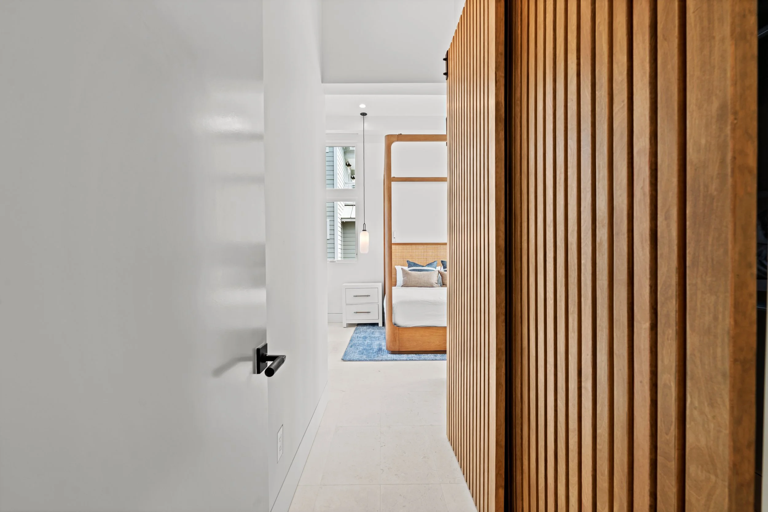 Modern bedroom with wooden bed frame, white walls, small white nightstand, blue rug, and window with exterior view